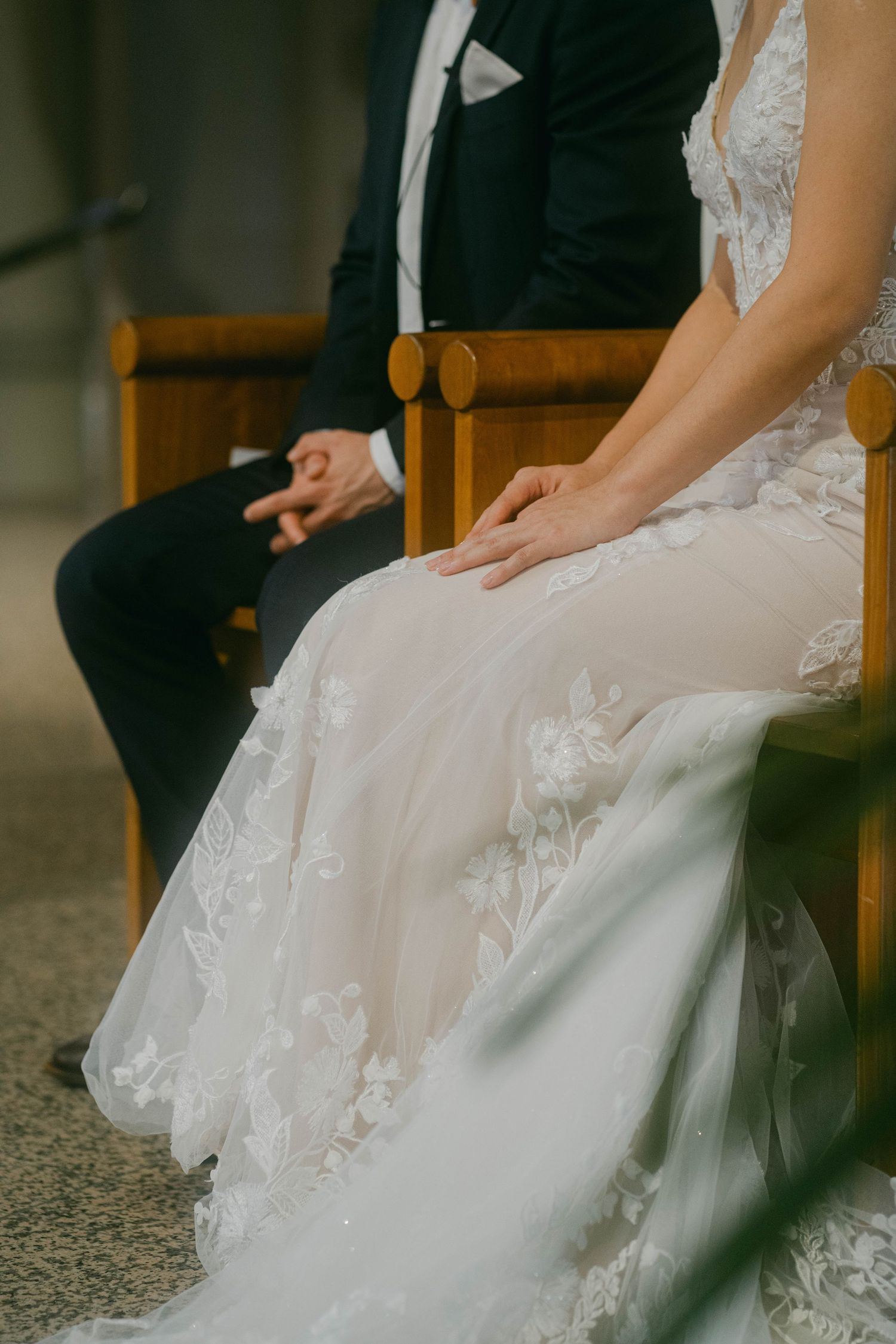 Detail shot of bride and groom sitting in wooden chairs, showing elegant white lace wedding dress and black suit.