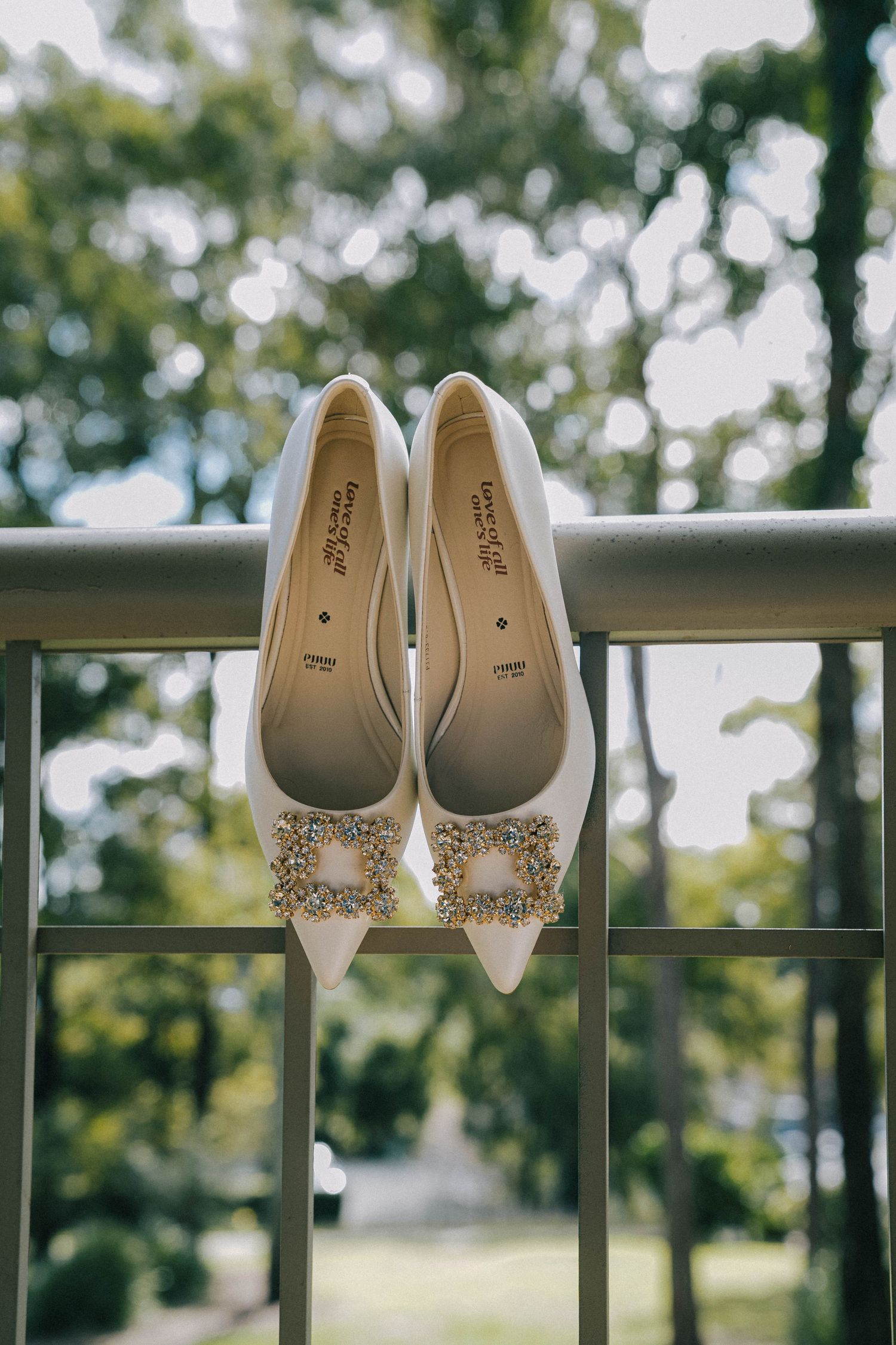 Elegant white bridal shoes with crystal embellishments displayed in a row on an outdoor railing with greenery in background.