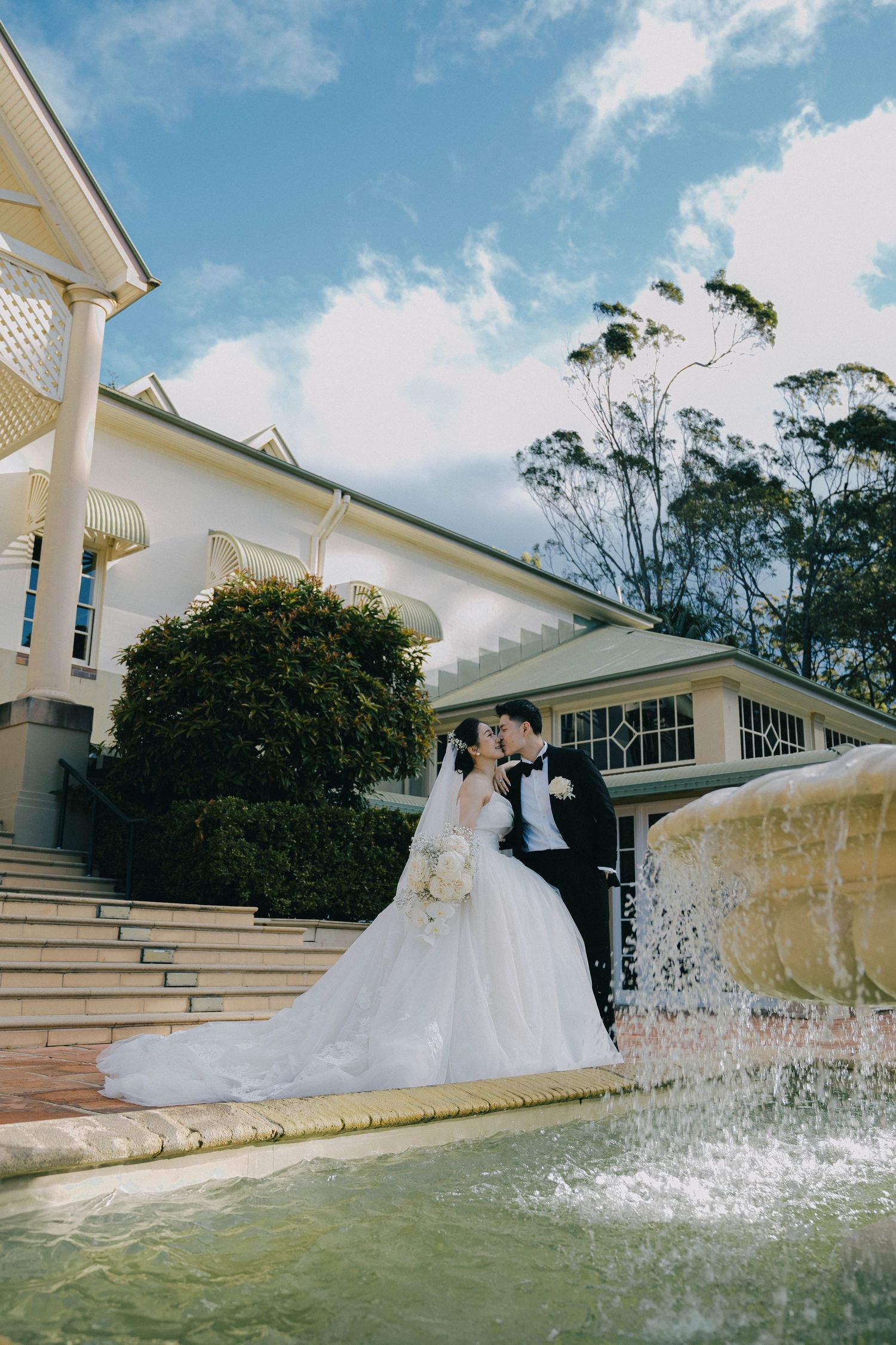 A wedding couple stands near a cascading water fountain at an elegant white mansion venue on a sunny day.