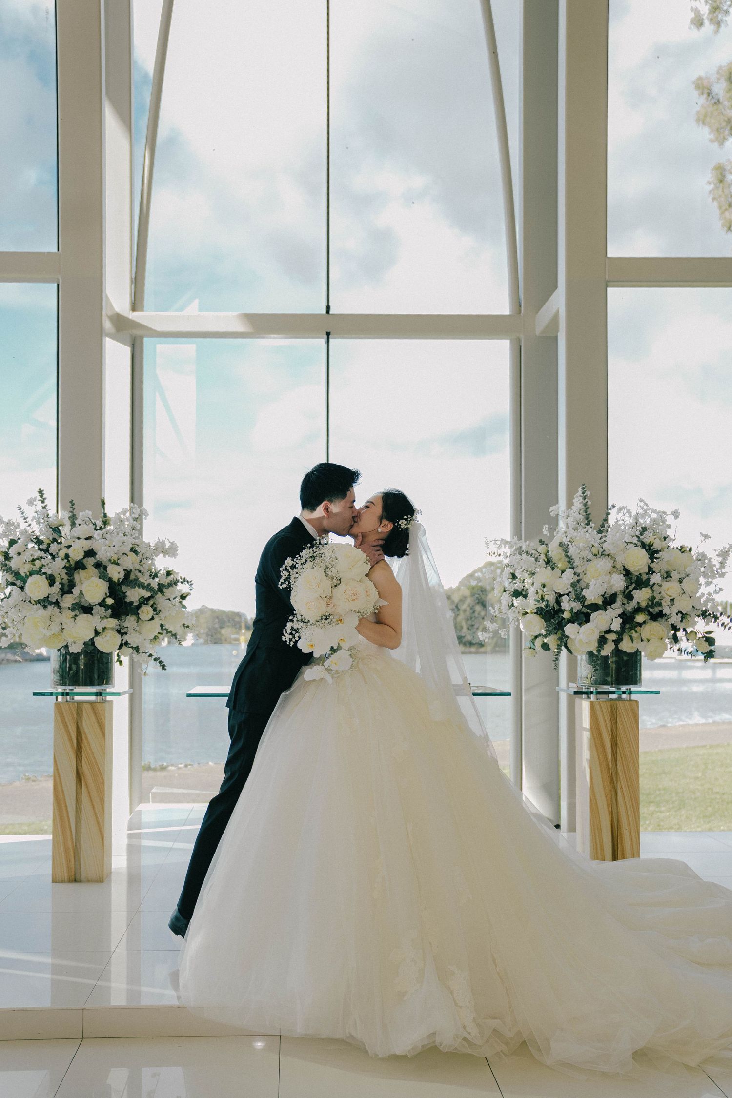 A romantic wedding couple shares a tender moment in a bright white chapel with floral arrangements and waterfront views.