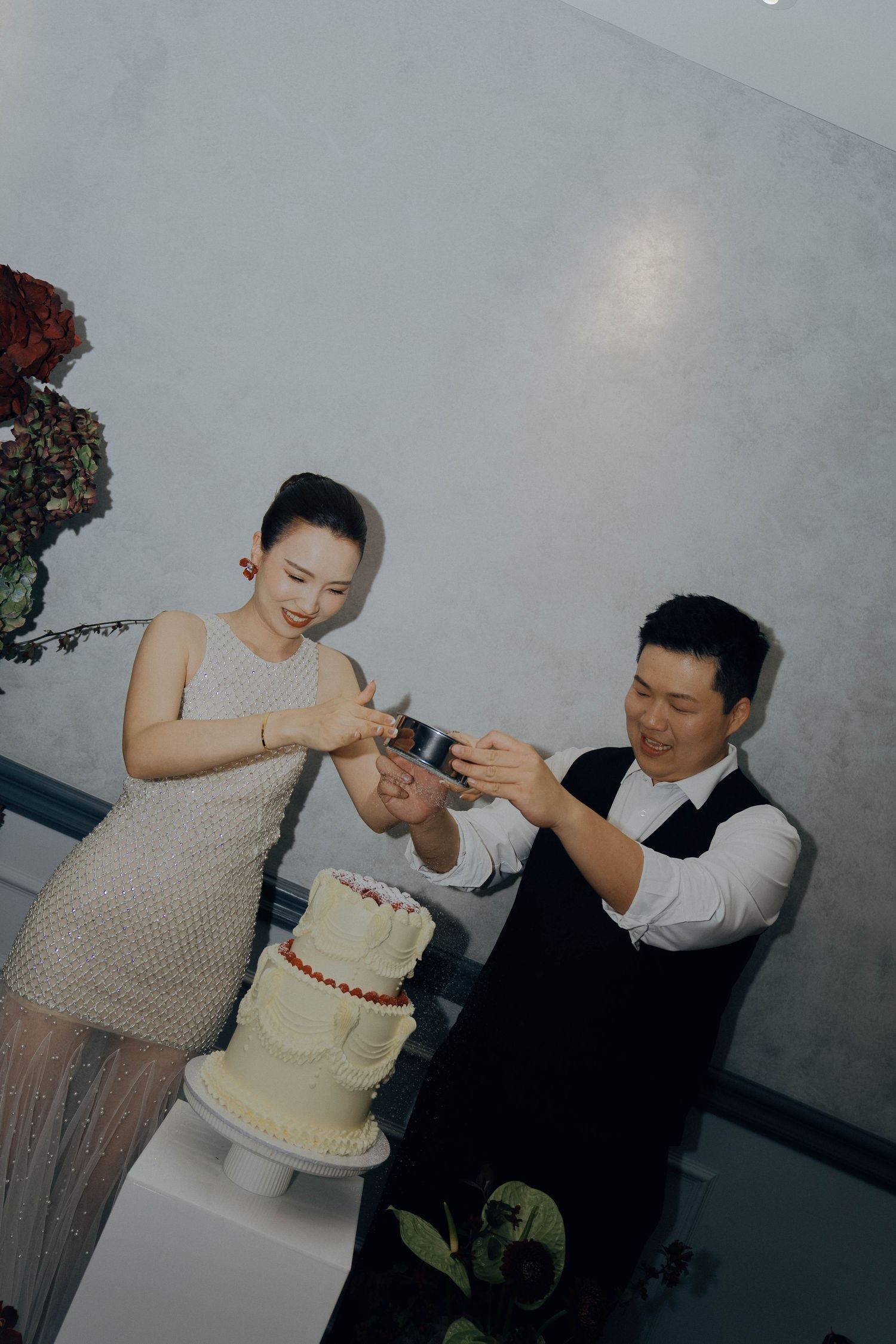 Two people in formal attire work together to decorate a white tiered cake with yellow trim in a studio setting.