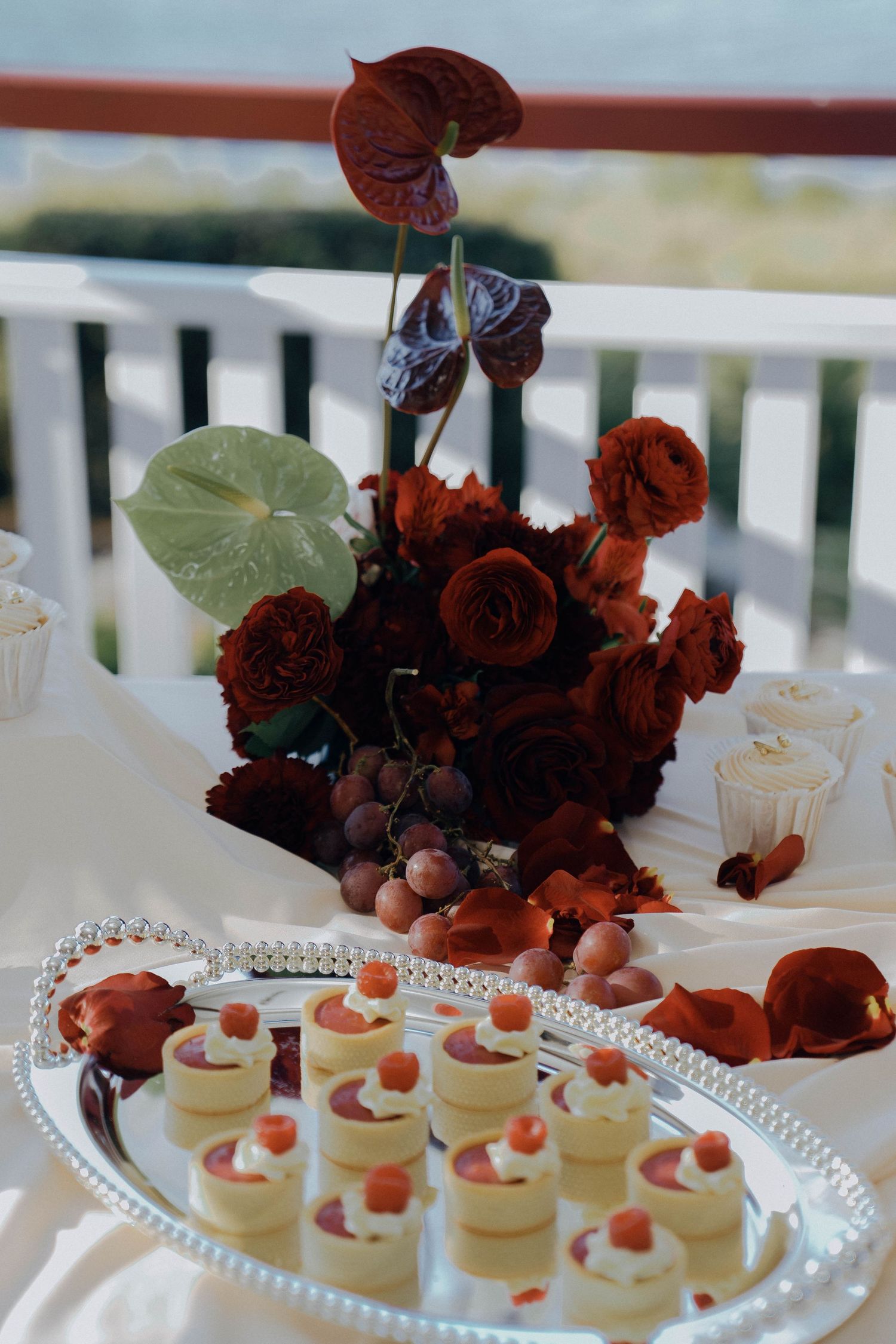 Elegant dessert display featuring white mini cheesecakes garnished with strawberries and dark red floral arrangements.