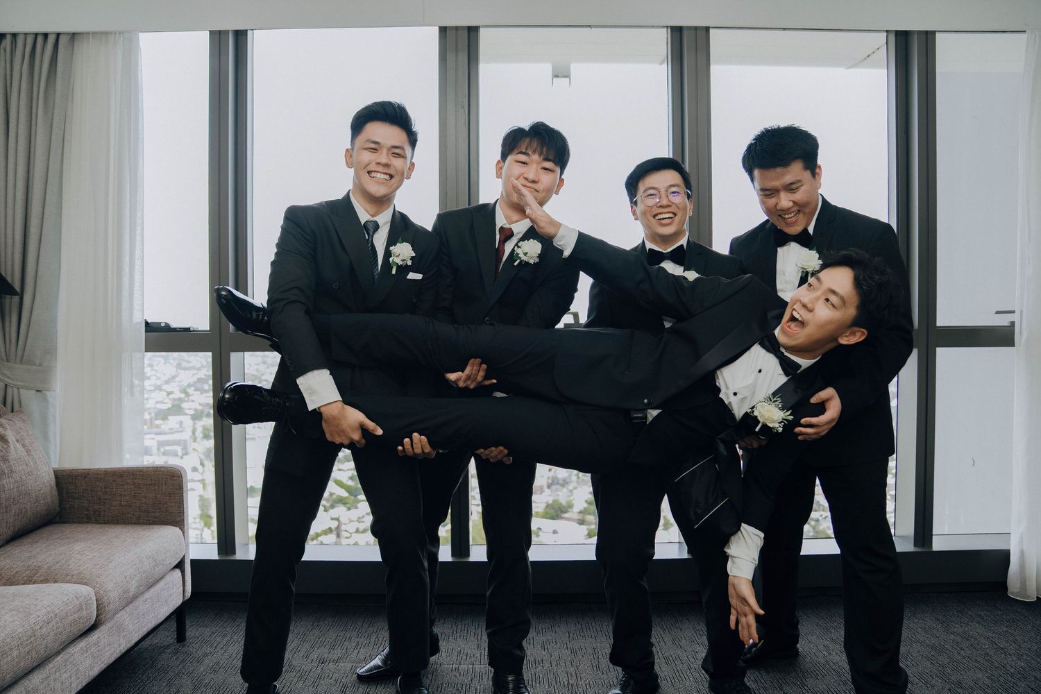 Groomsmen in black suits pose playfully together in a bright modern room for wedding photos.