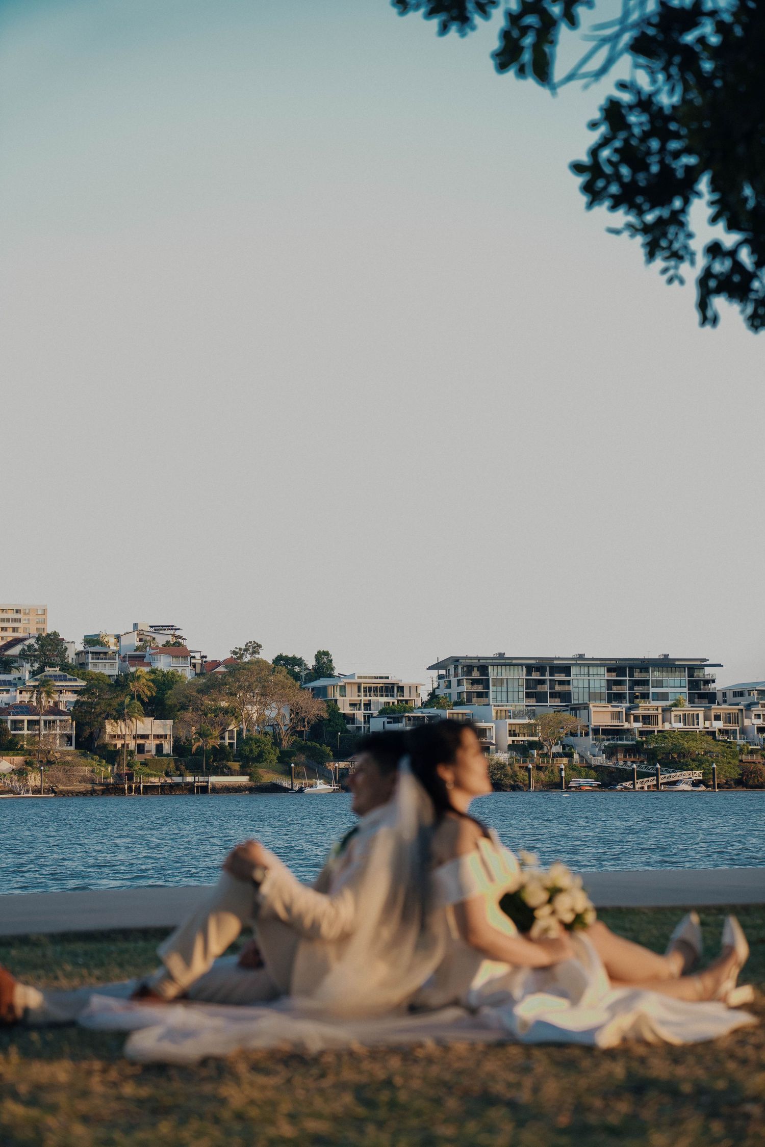 A couple relaxes on a blanket by a waterfront with buildings and trees visible in the background at sunset.