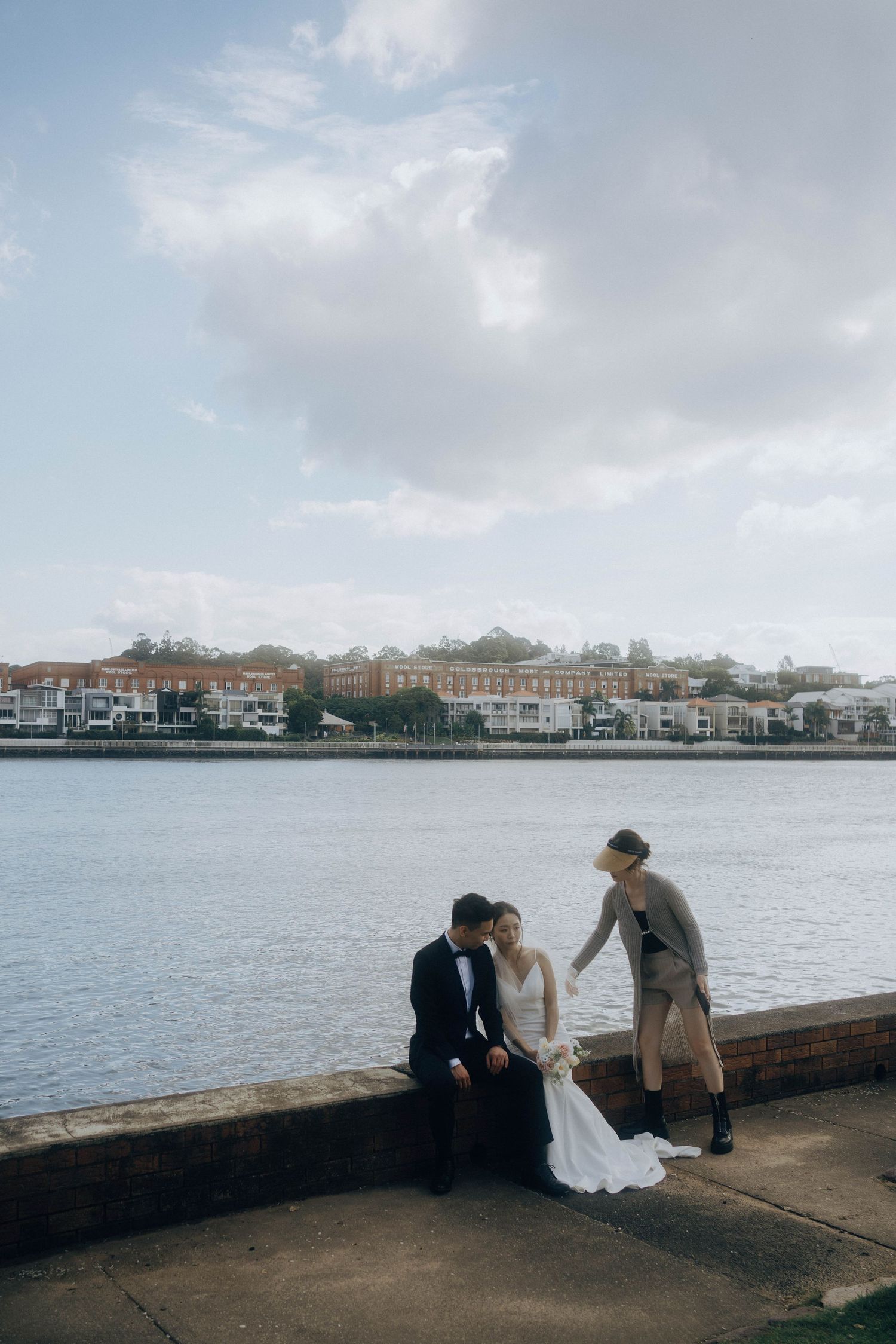 A couple sits on a waterfront pier with a scenic coastal town and cloudy sky in the background.
