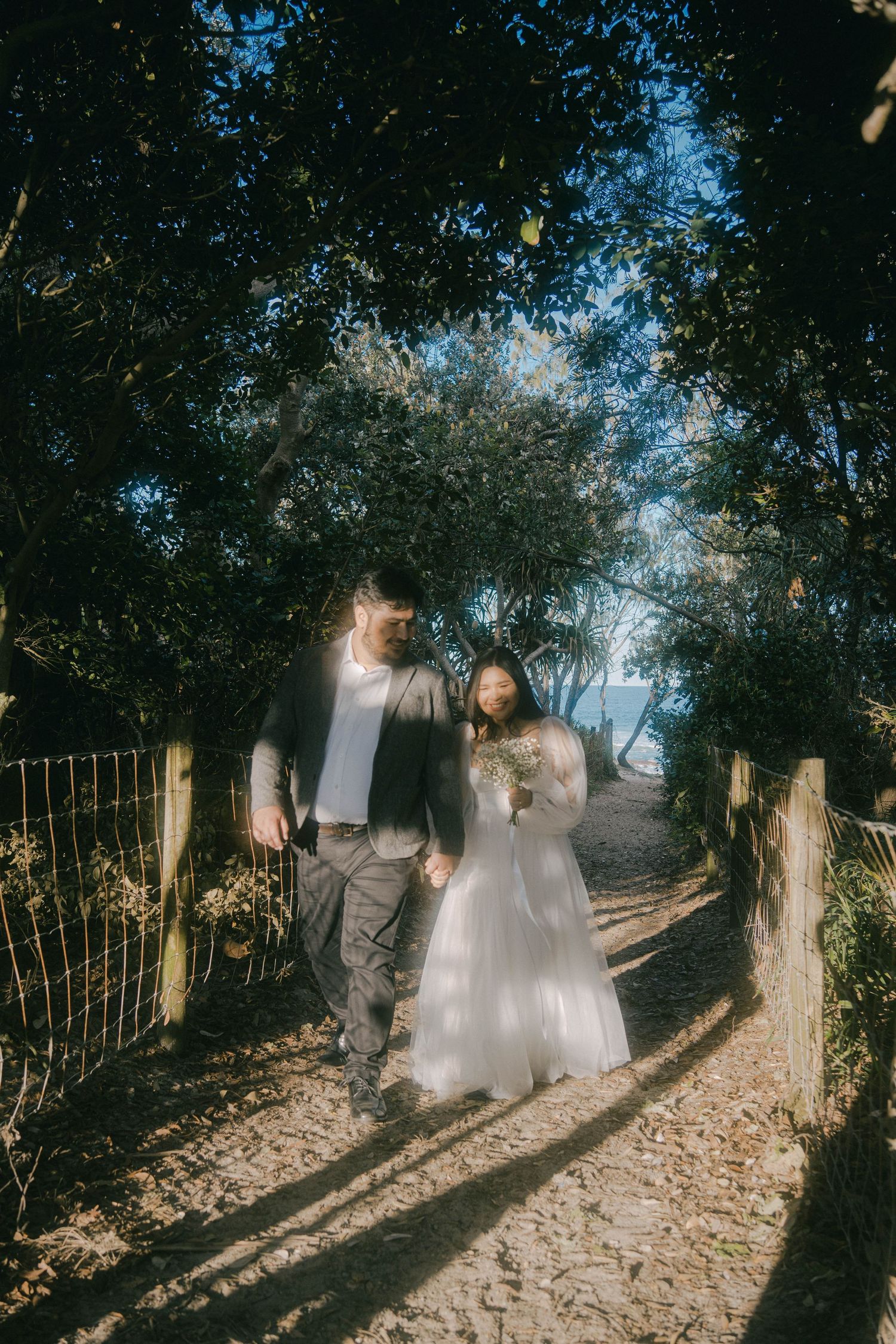 A couple walks along a sunlit forest path while shadows cast dramatic patterns through the trees.