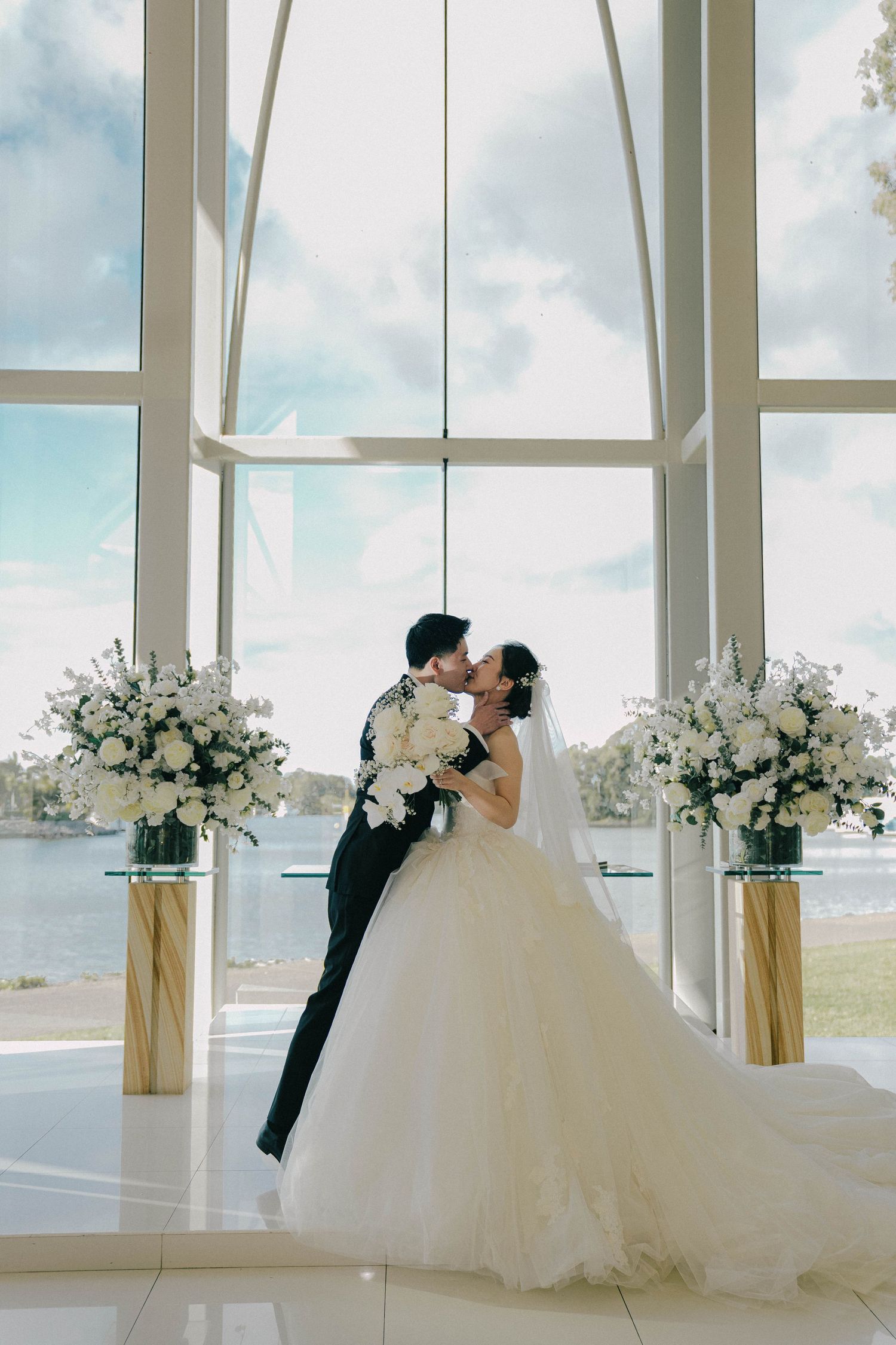 A romantic wedding couple shares a kiss in front of large windows overlooking the water, with elegant floral arrangements.