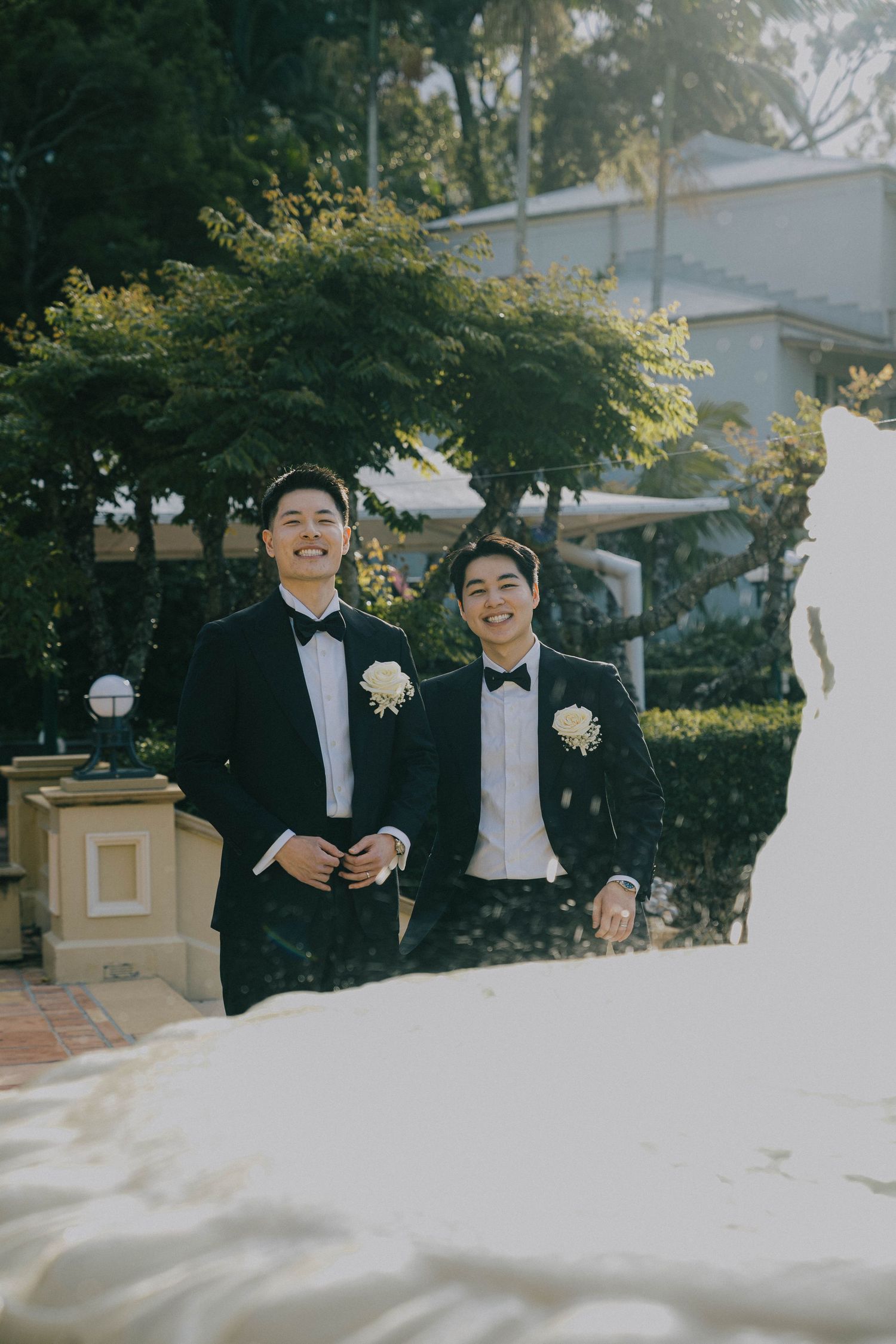 Two people in formal black tuxedos and bowties share a joyful moment outdoors during a special occasion.