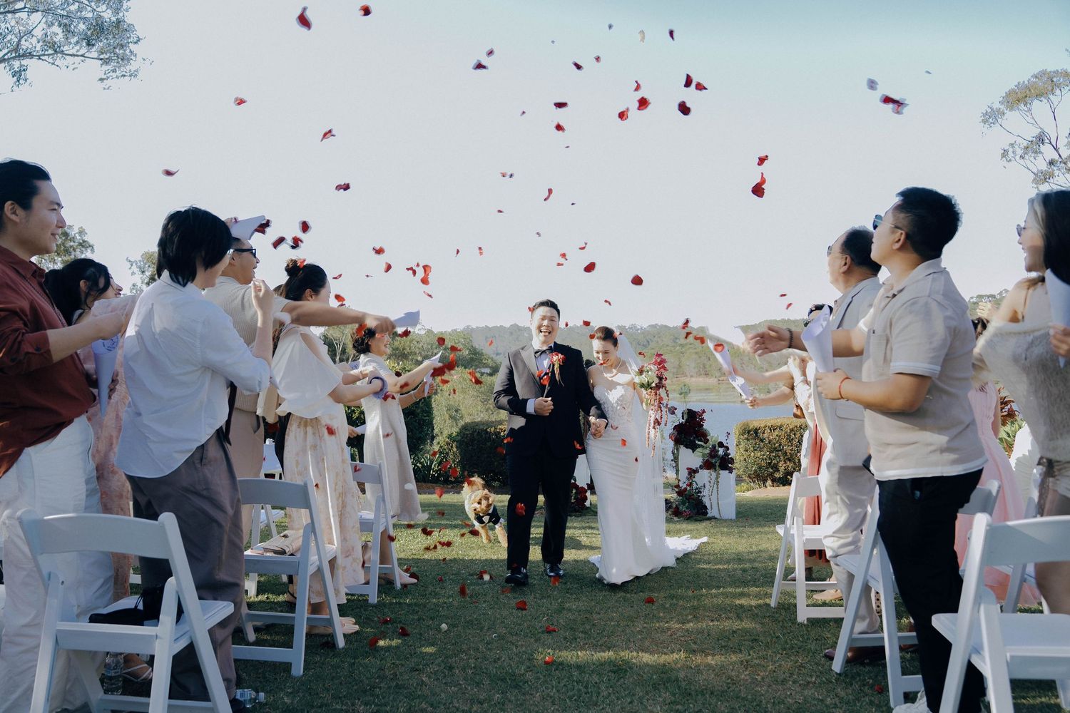 Wedding guests celebrate with red rose petals being tossed in the air during an outdoor garden ceremony.