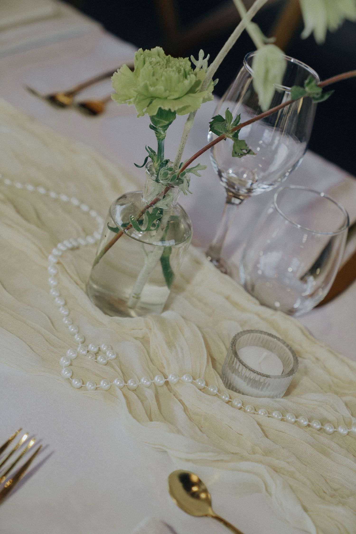 Elegant table setting with cream linens, pearl necklace decor, and delicate green floral arrangements in glass vases.
