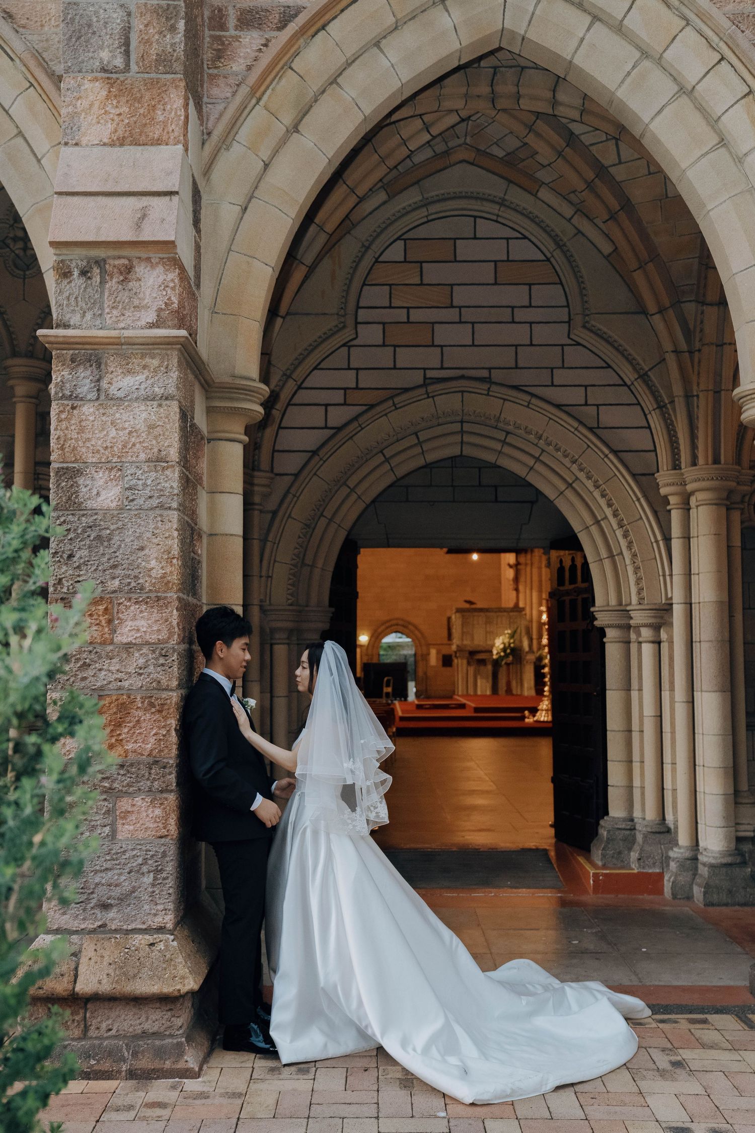 A couple stands beneath gothic stone archways at a church entrance, with the bride wearing a flowing white gown with long train.