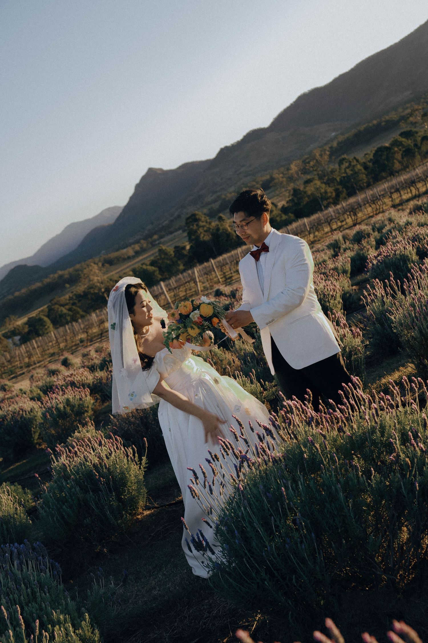 A couple in wedding attire dances together in a scenic mountain field with lavender flowers at sunset.