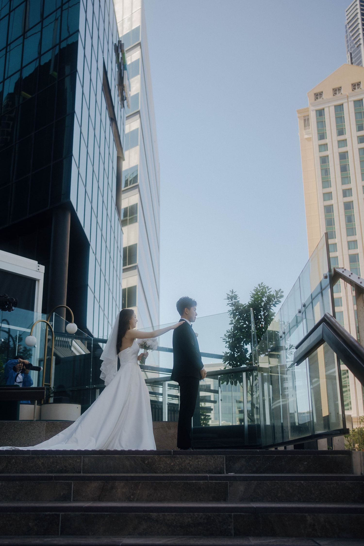 A couple in wedding attire stands on steps outside a modern glass office building on a sunny day.