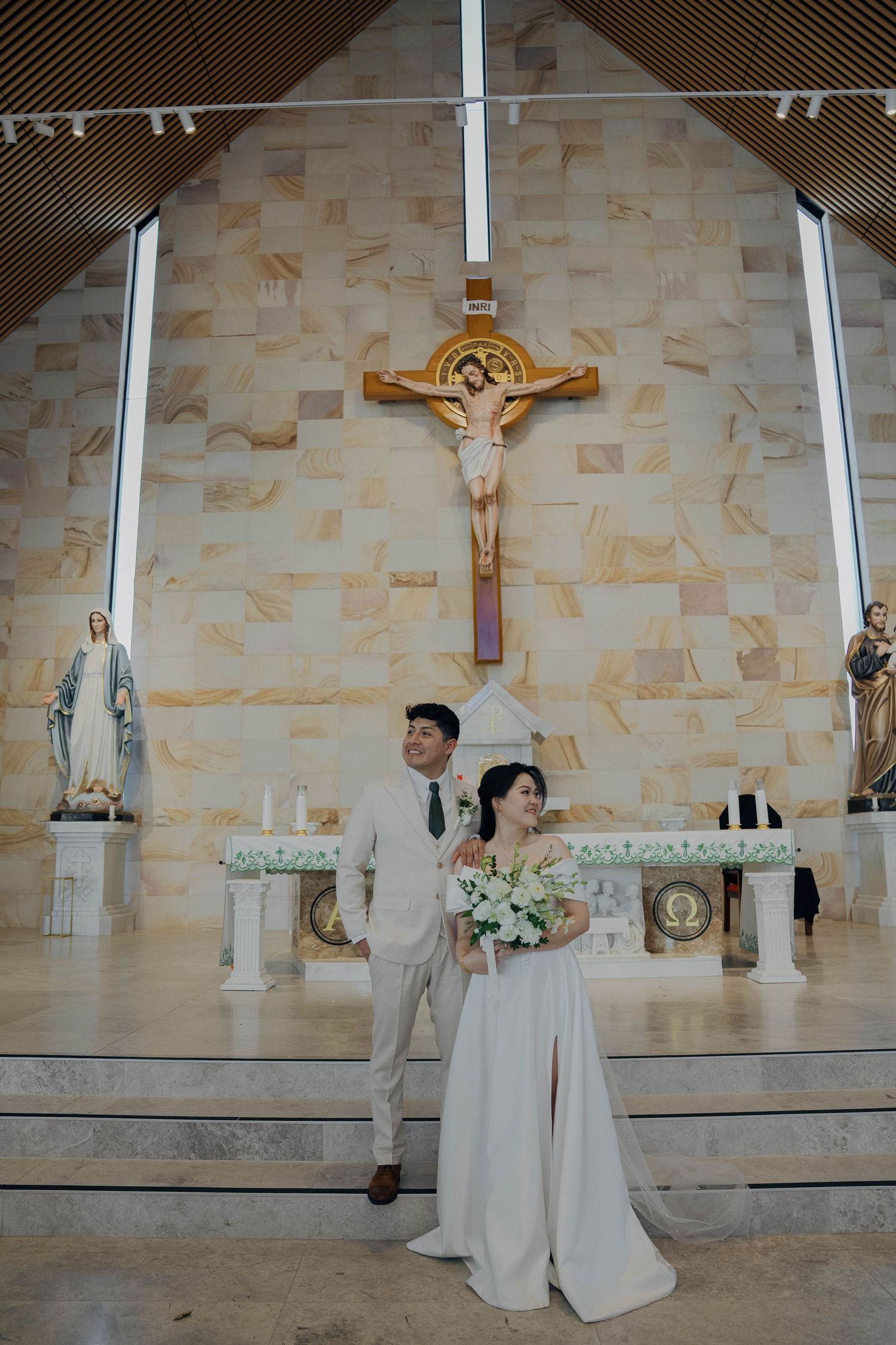 Wedding couple stands at church altar with stone wall backdrop and religious symbols including crucifixes and statues.