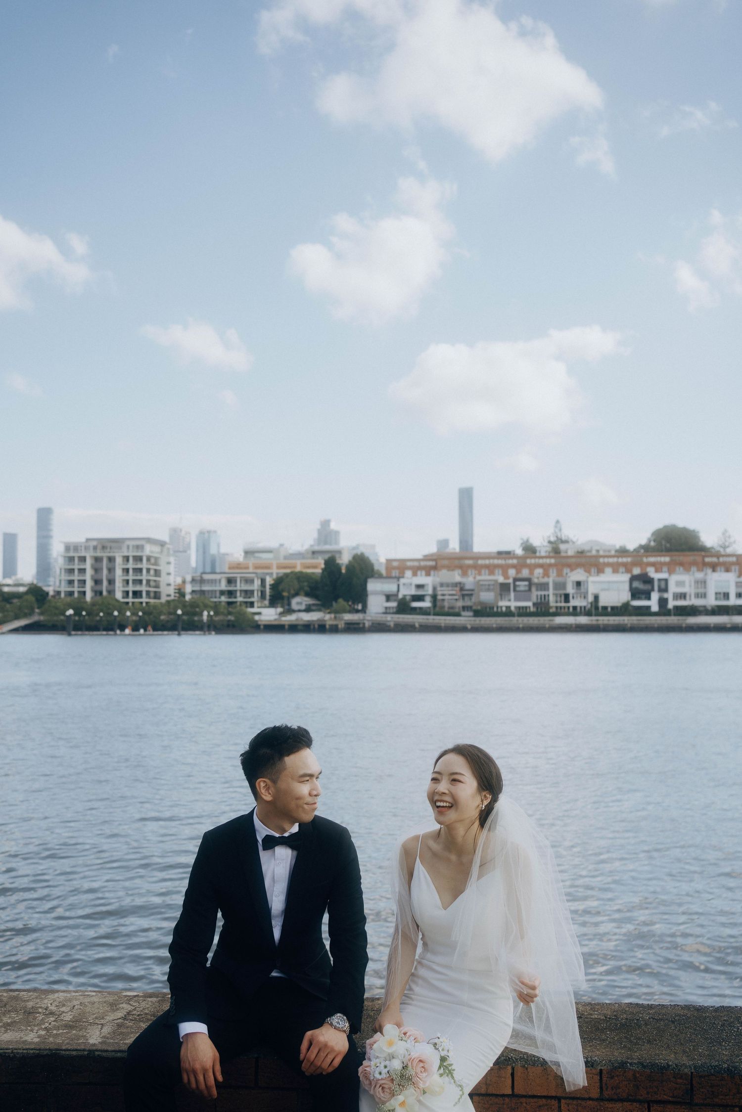 A couple sits by the waterfront with a cityscape backdrop in Singapore during their wedding photoshoot.