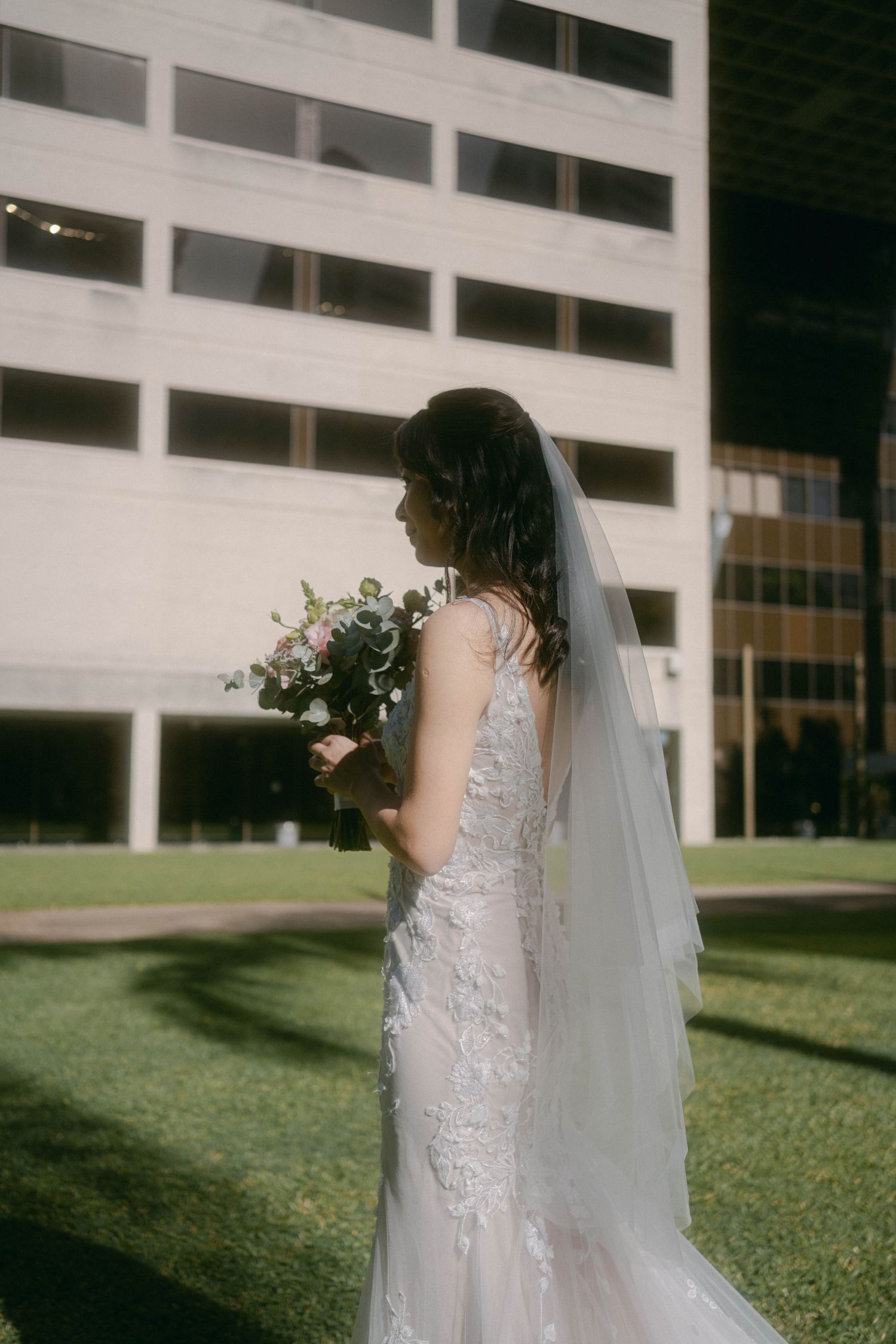 A bride in a detailed lace wedding gown and long veil holds a bouquet while standing on grass near a modern building.