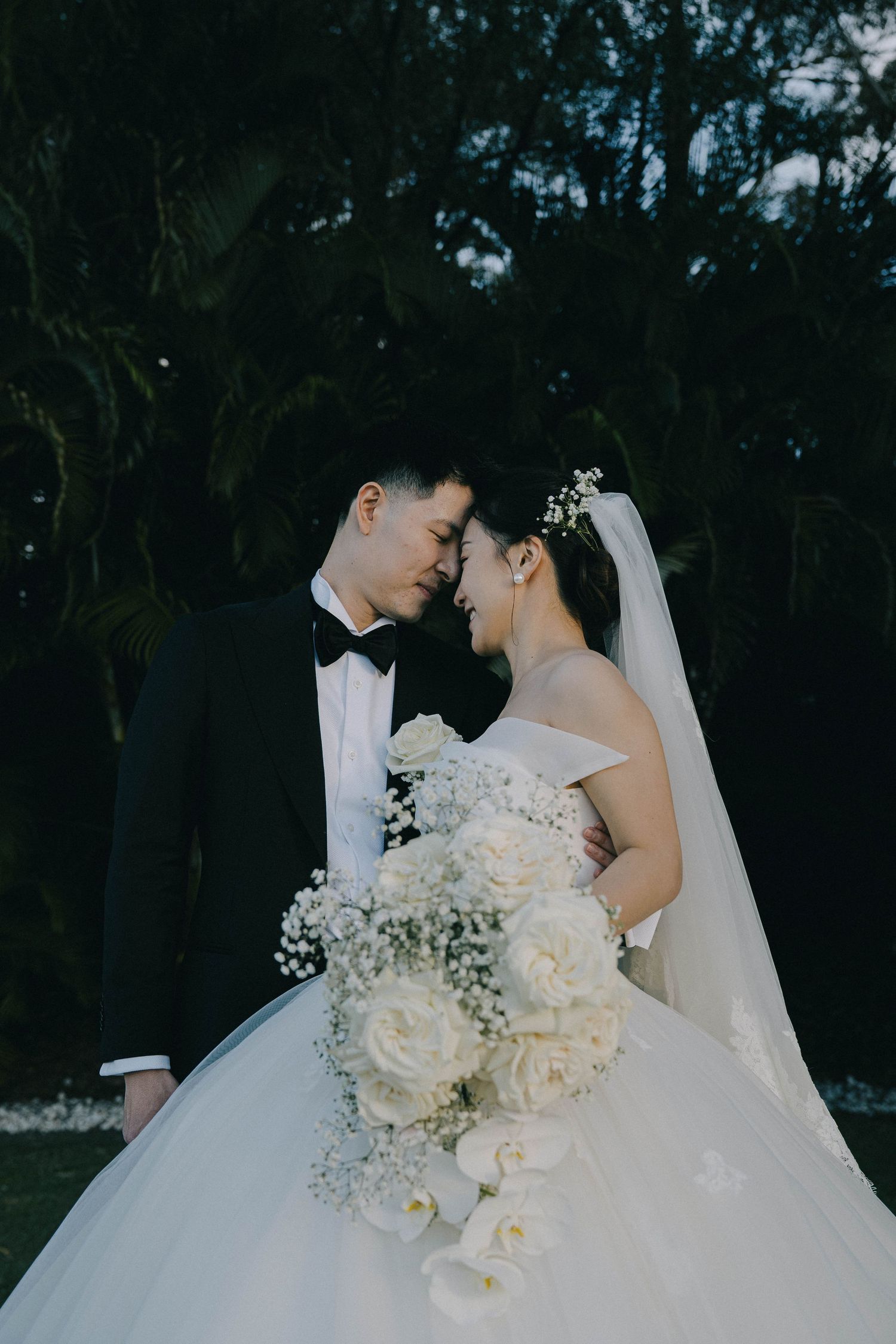 A romantic wedding couple embraces outdoors at dusk with a beautiful white floral bouquet.