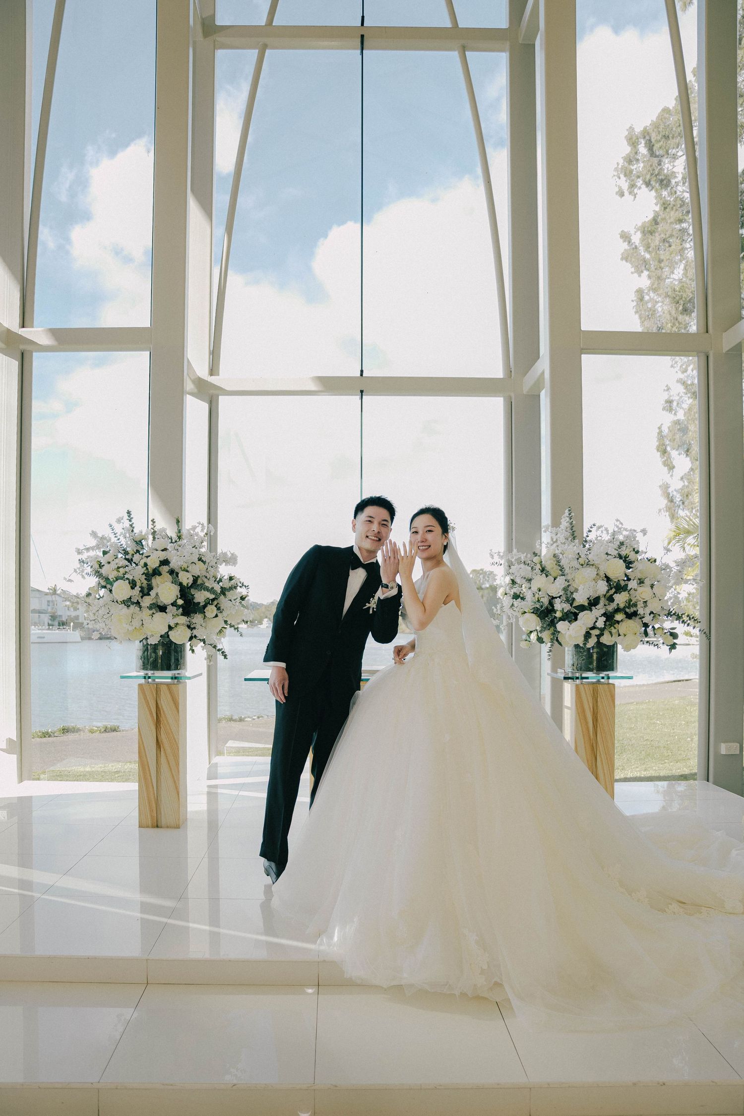 A couple in wedding attire poses in a bright white chapel with floor-to-ceiling windows and floral arrangements.