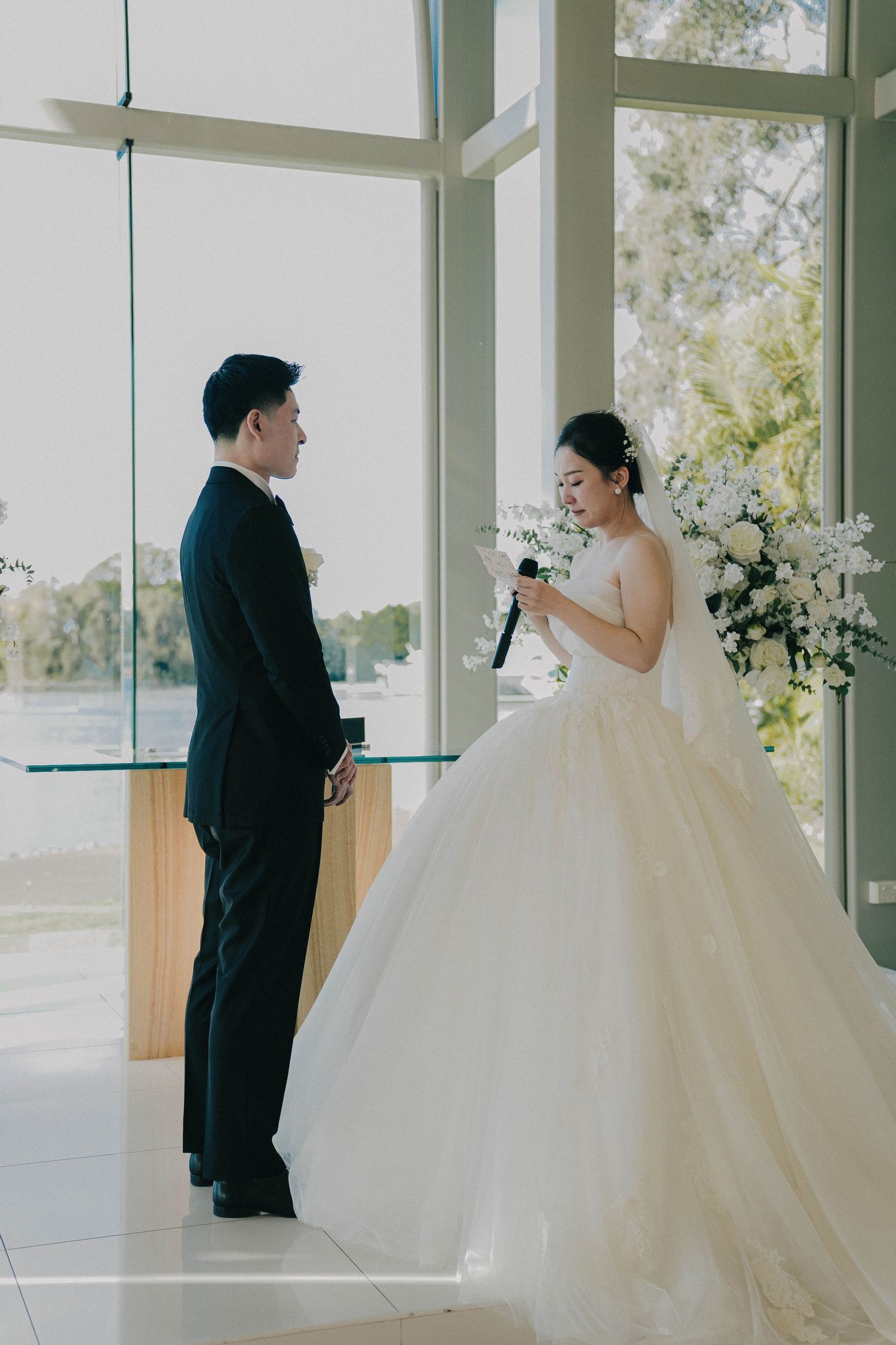 A couple stands together in a bright chapel with floor-to-ceiling windows and white floral arrangements.
