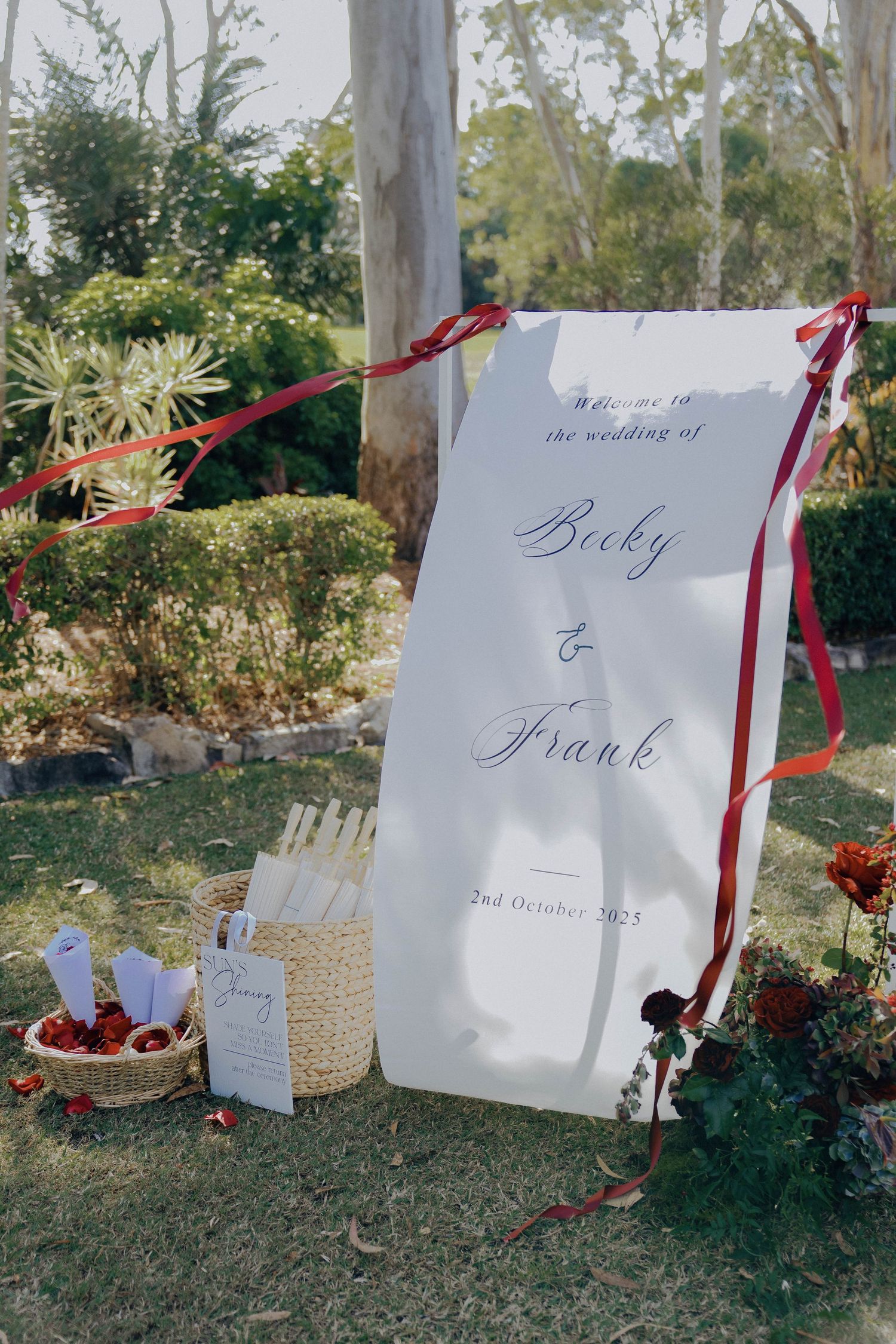 Elegant wedding welcome sign with red ribbons displayed in garden setting next to refreshments and decor.