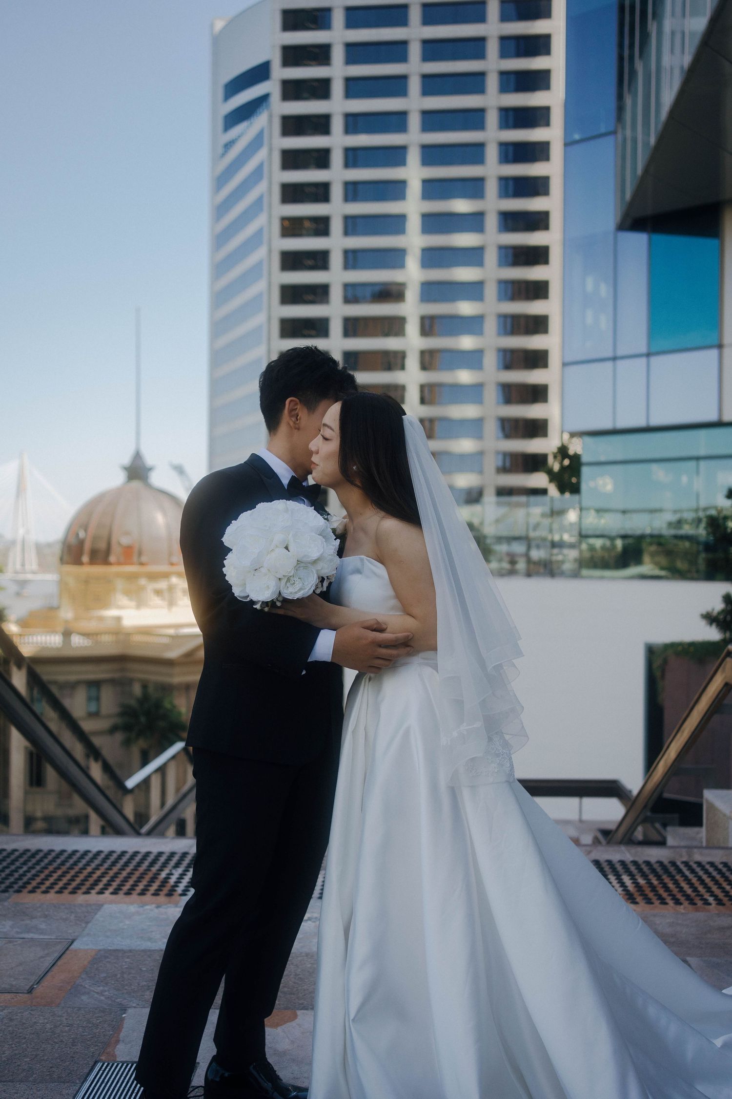 A romantic wedding couple embraces on an urban rooftop with modern buildings in the background.