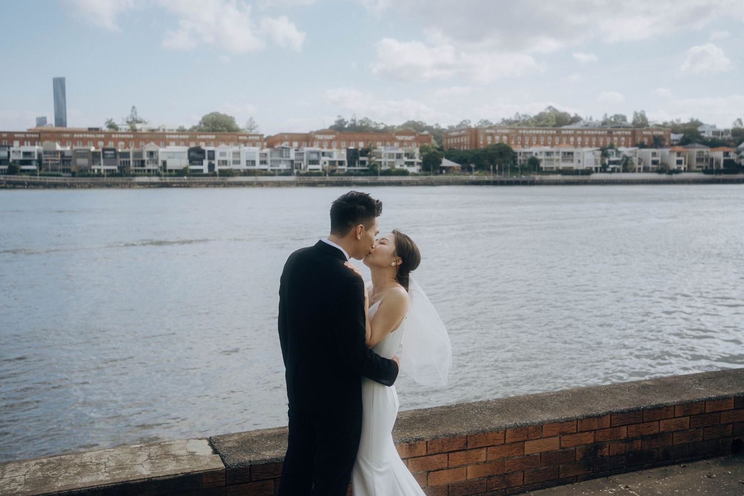A couple embraces during their wedding photoshoot along a waterfront with boats and buildings in the background.