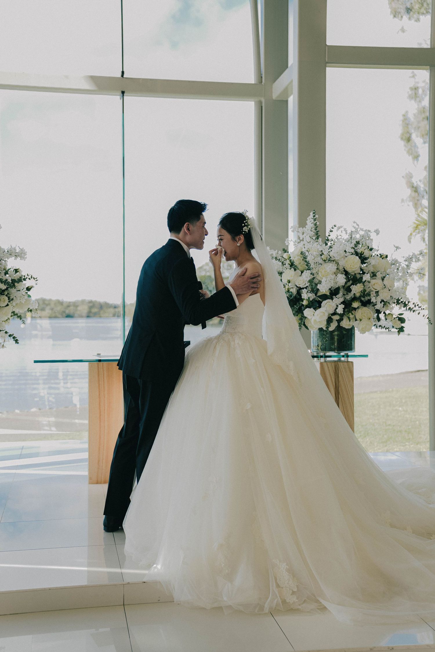 A couple shares an intimate moment in a modern chapel with floor-to-ceiling windows and white floral arrangements.