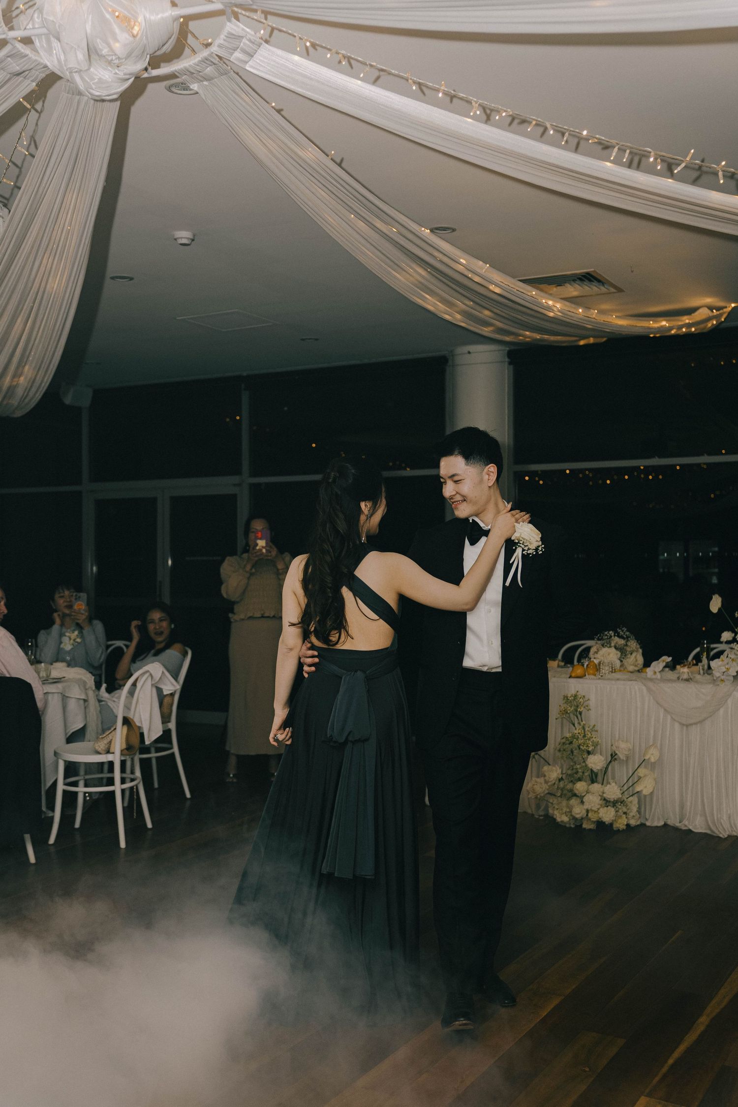 A couple dances together on a dark dance floor with atmospheric fog during what appears to be a wedding reception.
