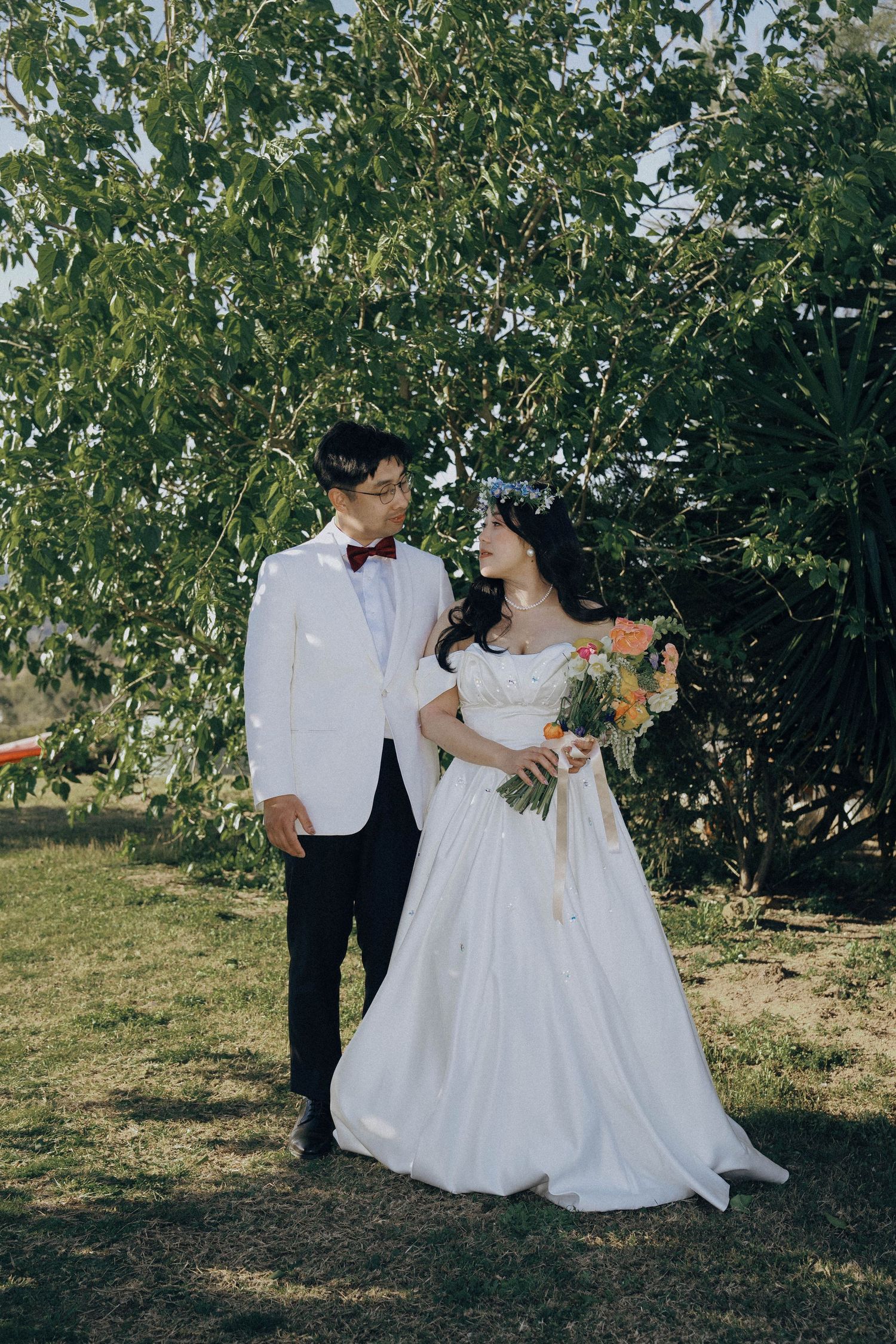 A couple in wedding attire shares romantic moments in a sunny garden setting with green trees in the background.