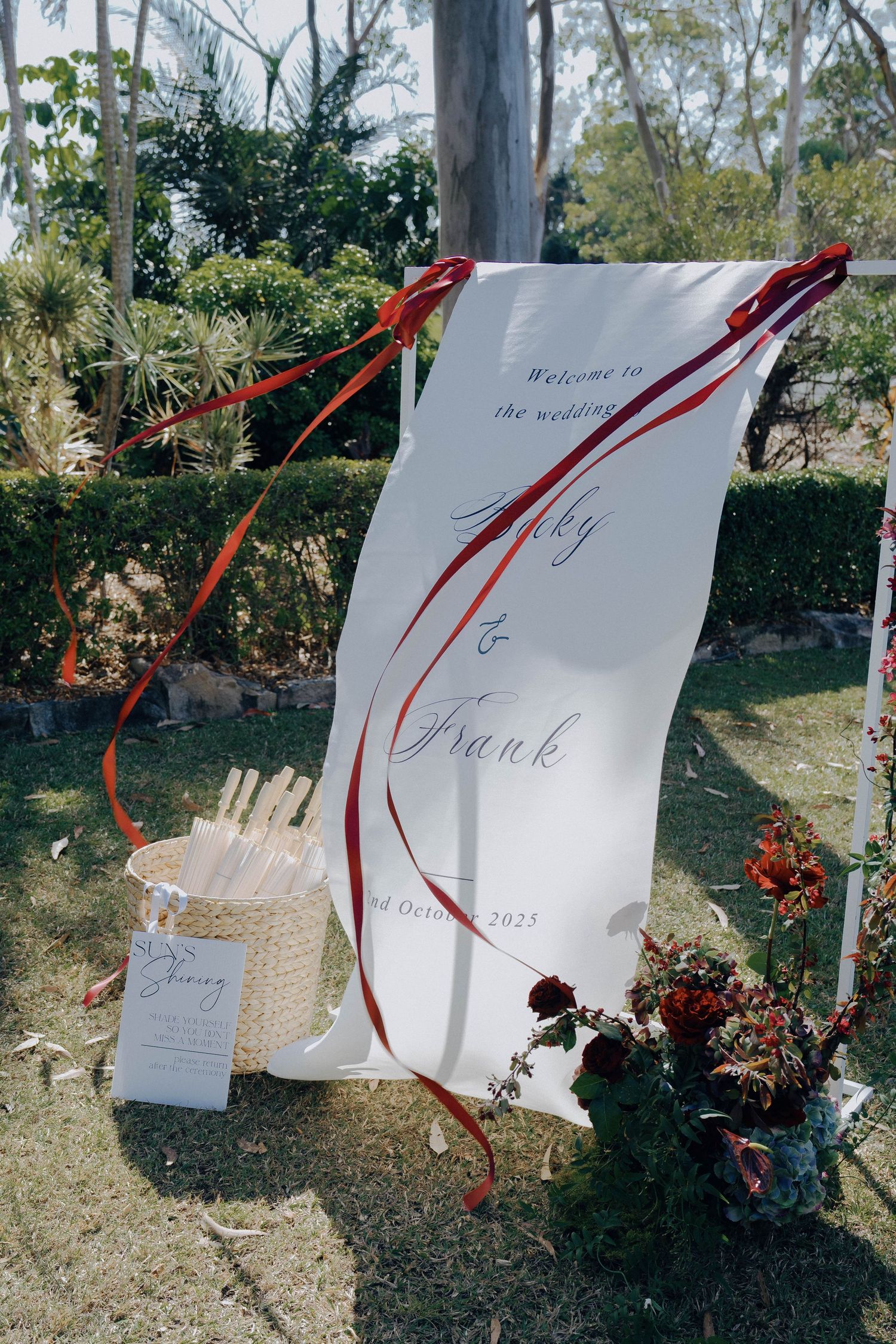 White paper wedding invitations with red ribbons hang on display in an outdoor garden setting.