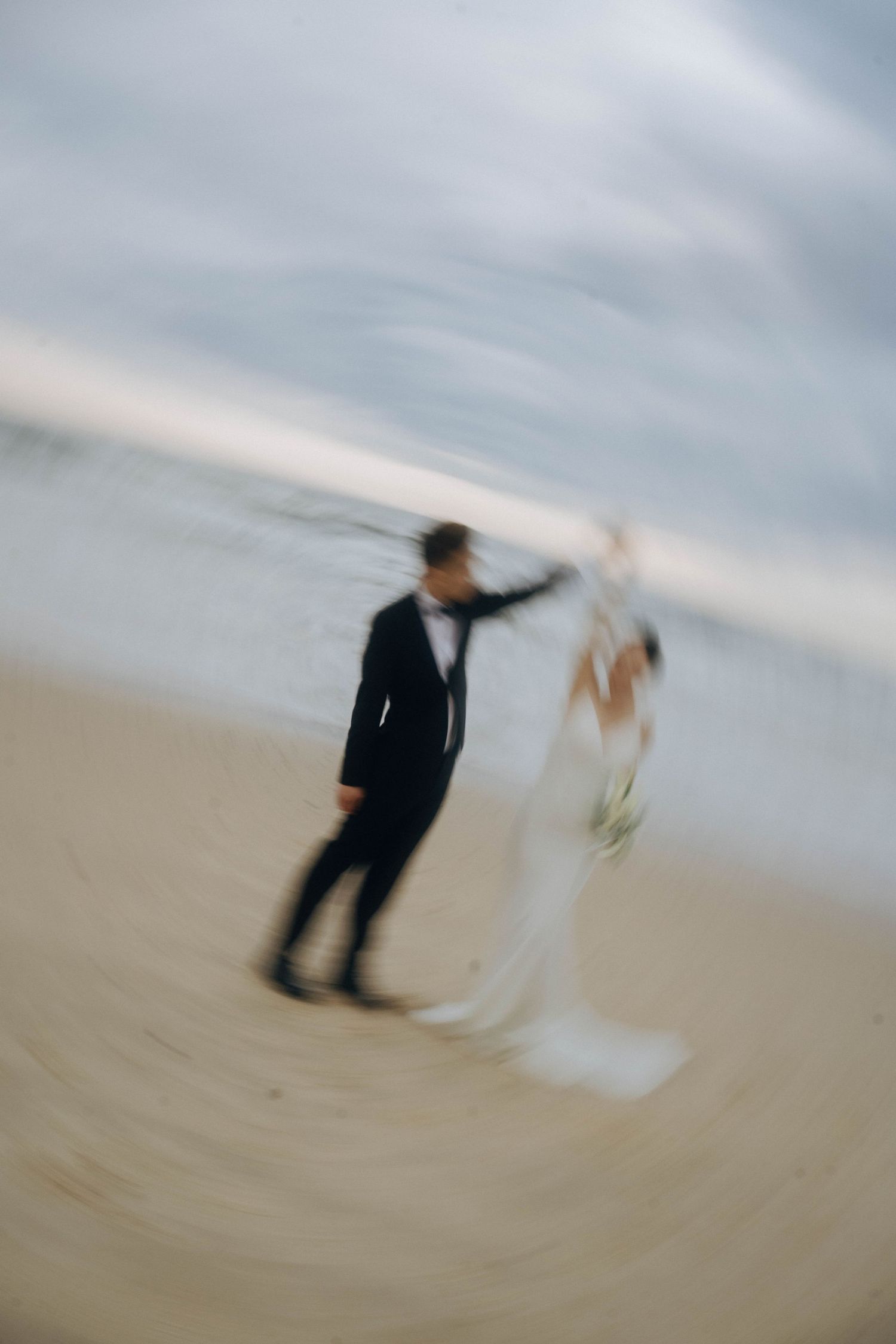 A couple in wedding attire walking along a misty beach shore captured in an artistic blurred motion effect.