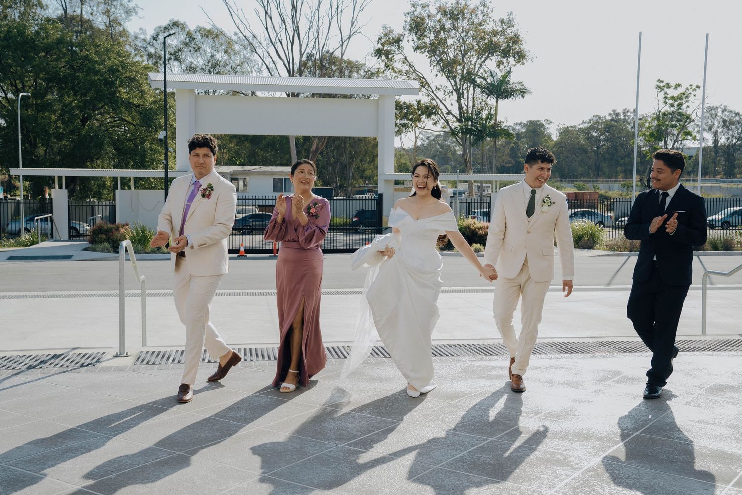 Wedding party members walk and dance in a line formation on a sunny outdoor pathway casting long shadows.
