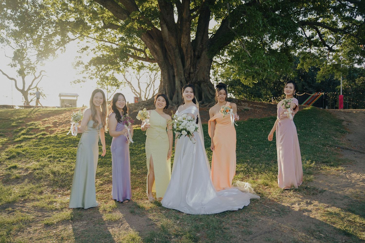 A wedding party in pastel dresses stands together on a grassy hillside during golden hour sunset.