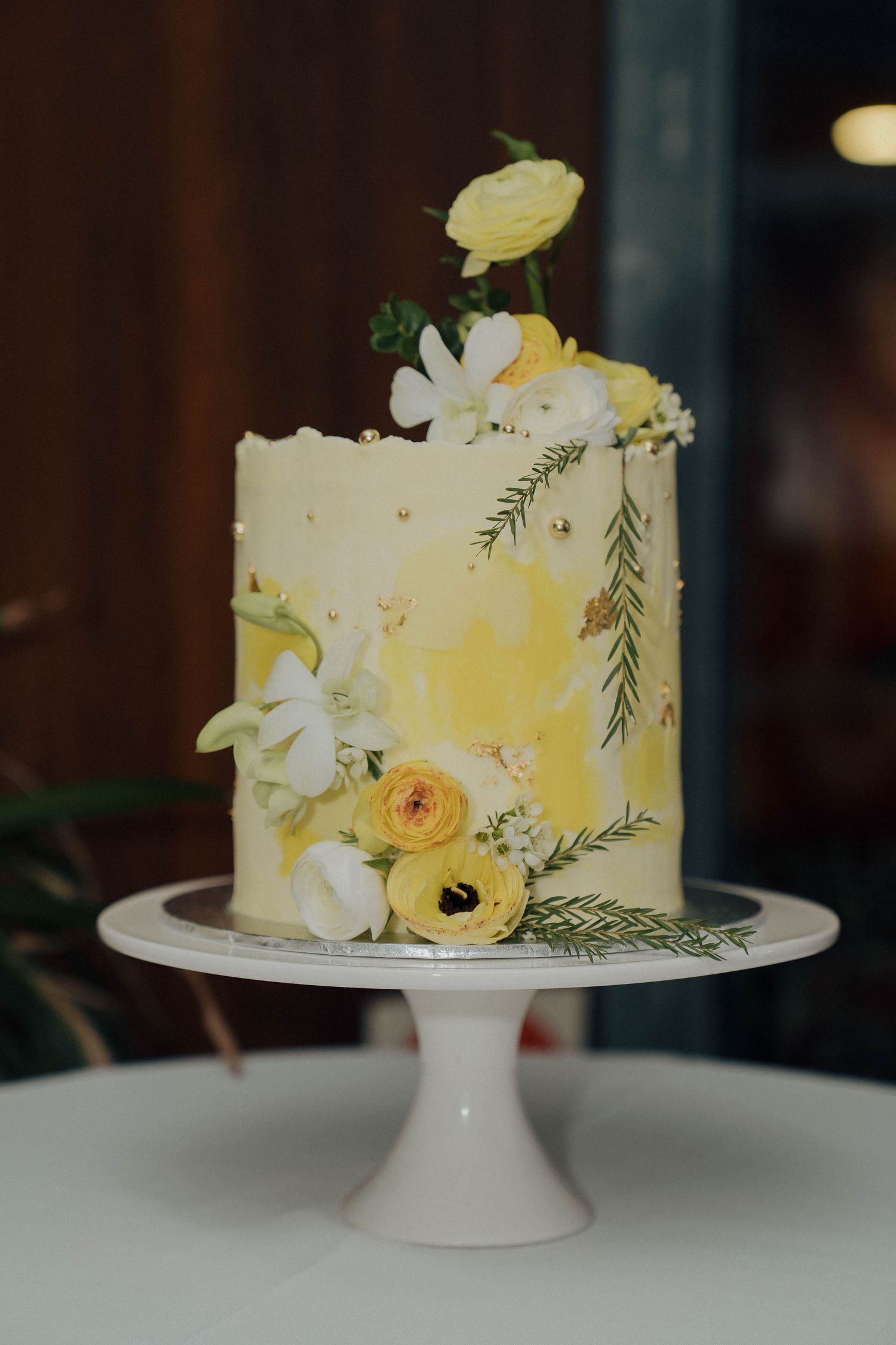 Elegant white and yellow wedding cake decorated with fresh ranunculus flowers and greenery on a white pedestal stand.
