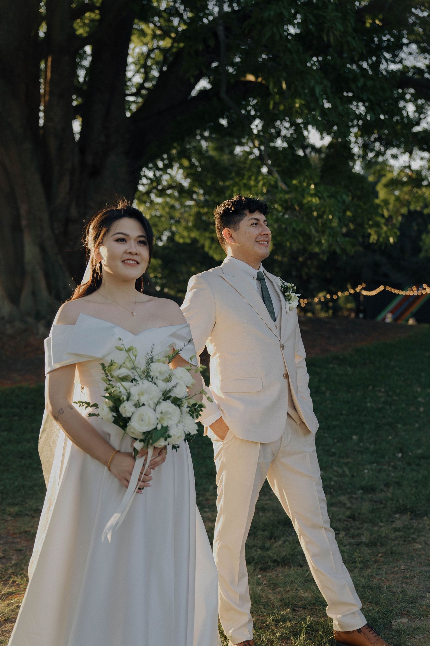 A couple in white wedding attire stands together outdoors with a bouquet of white flowers at sunset.