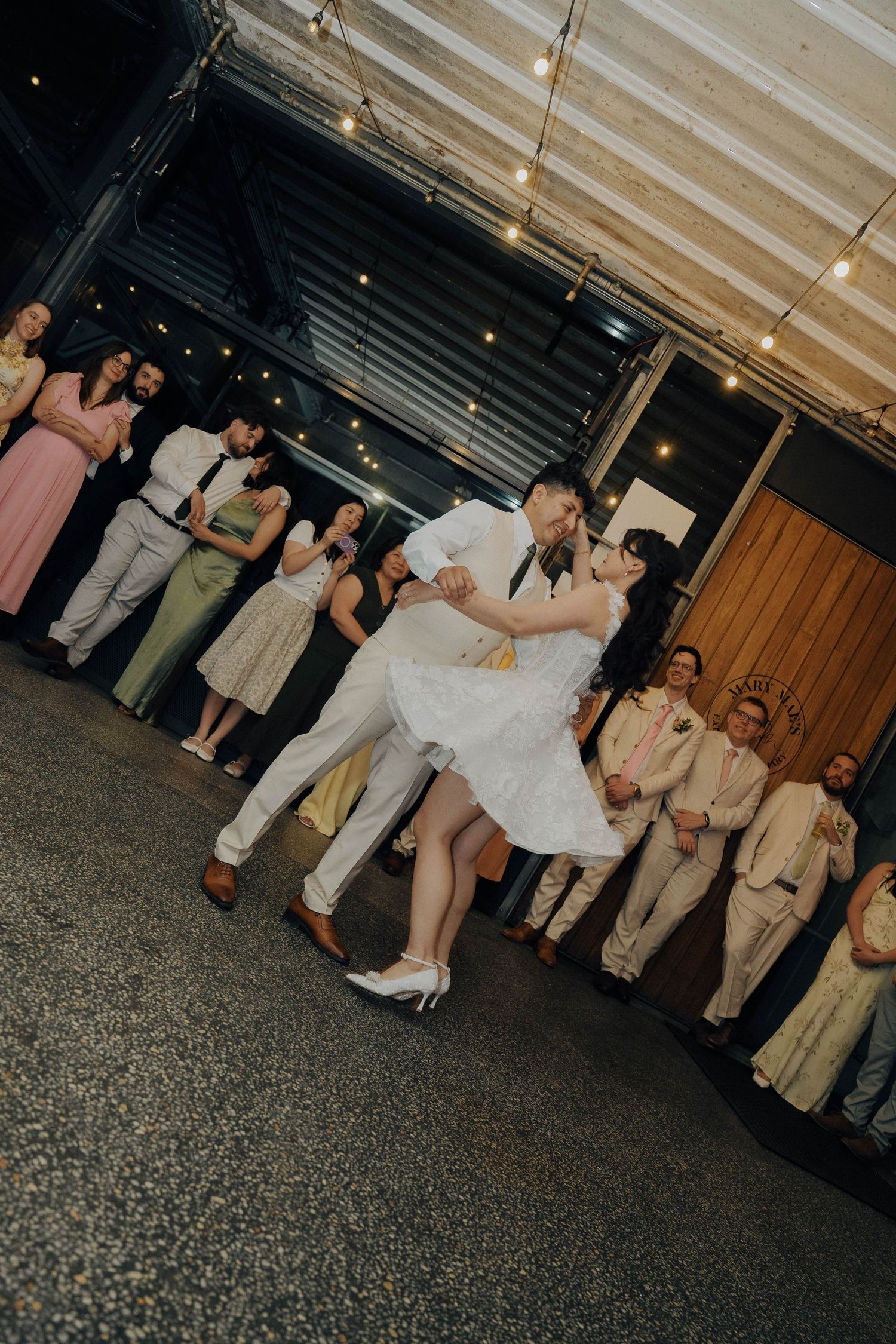 A couple performs a romantic dip dance move at their wedding reception while guests look on under string lights.