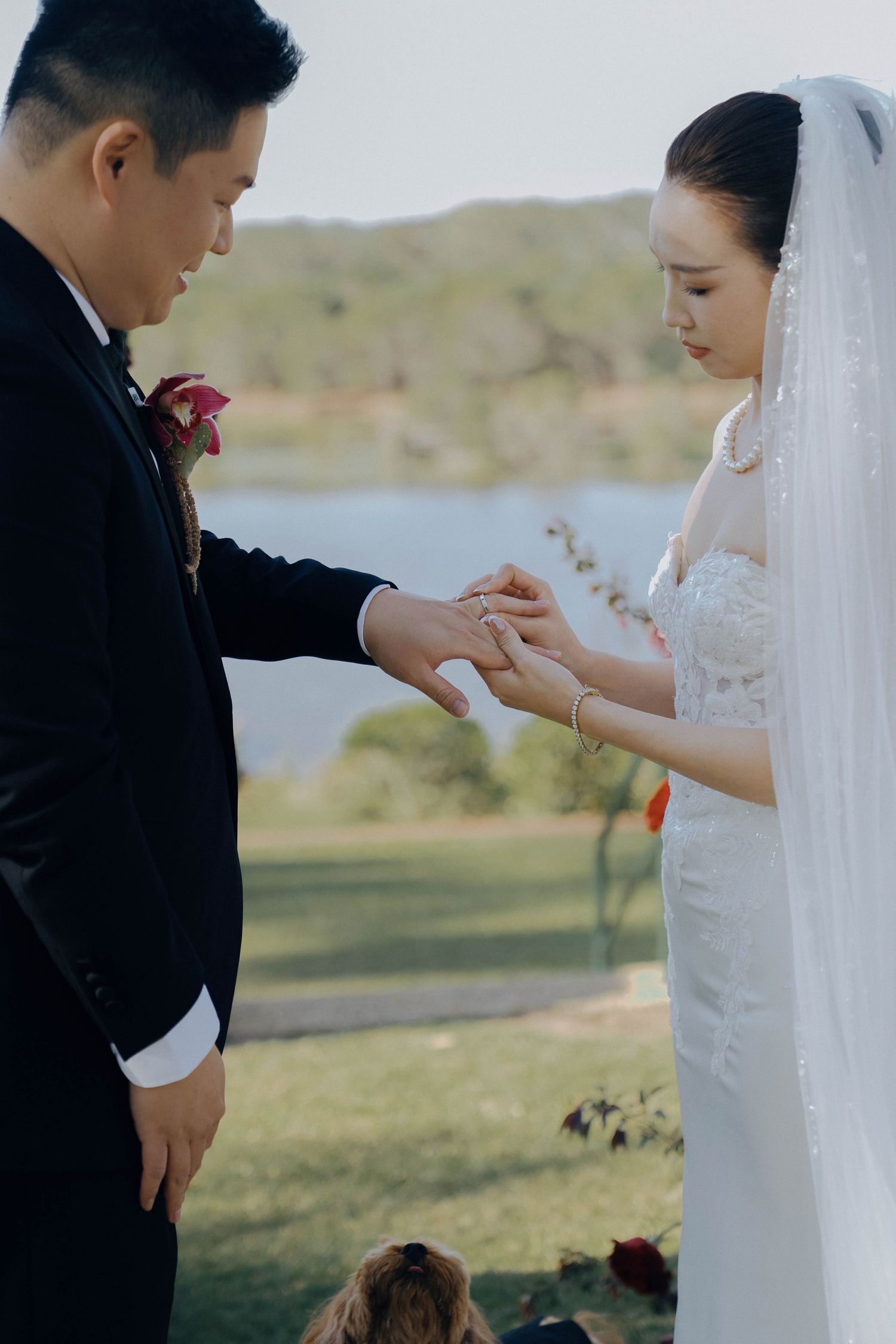 A wedding couple exchanges rings during an outdoor ceremony on a grassy hillside.