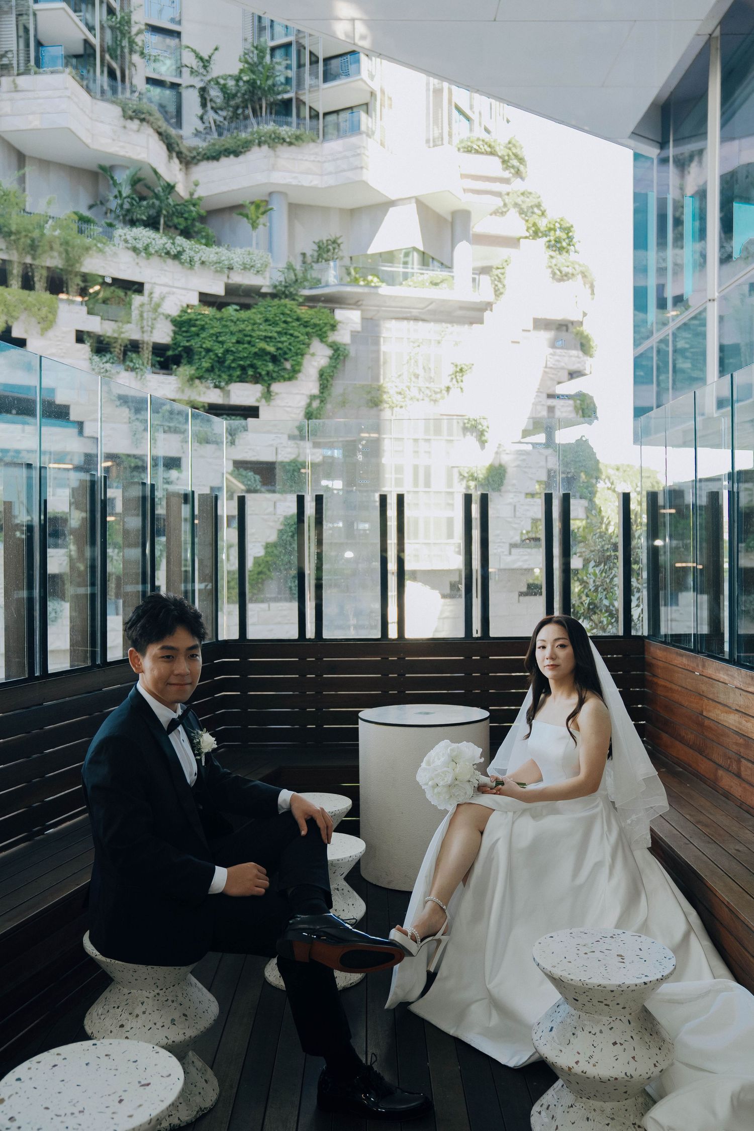 A couple in wedding attire sits on a modern bench in a bright, glass-enclosed space with architectural details.