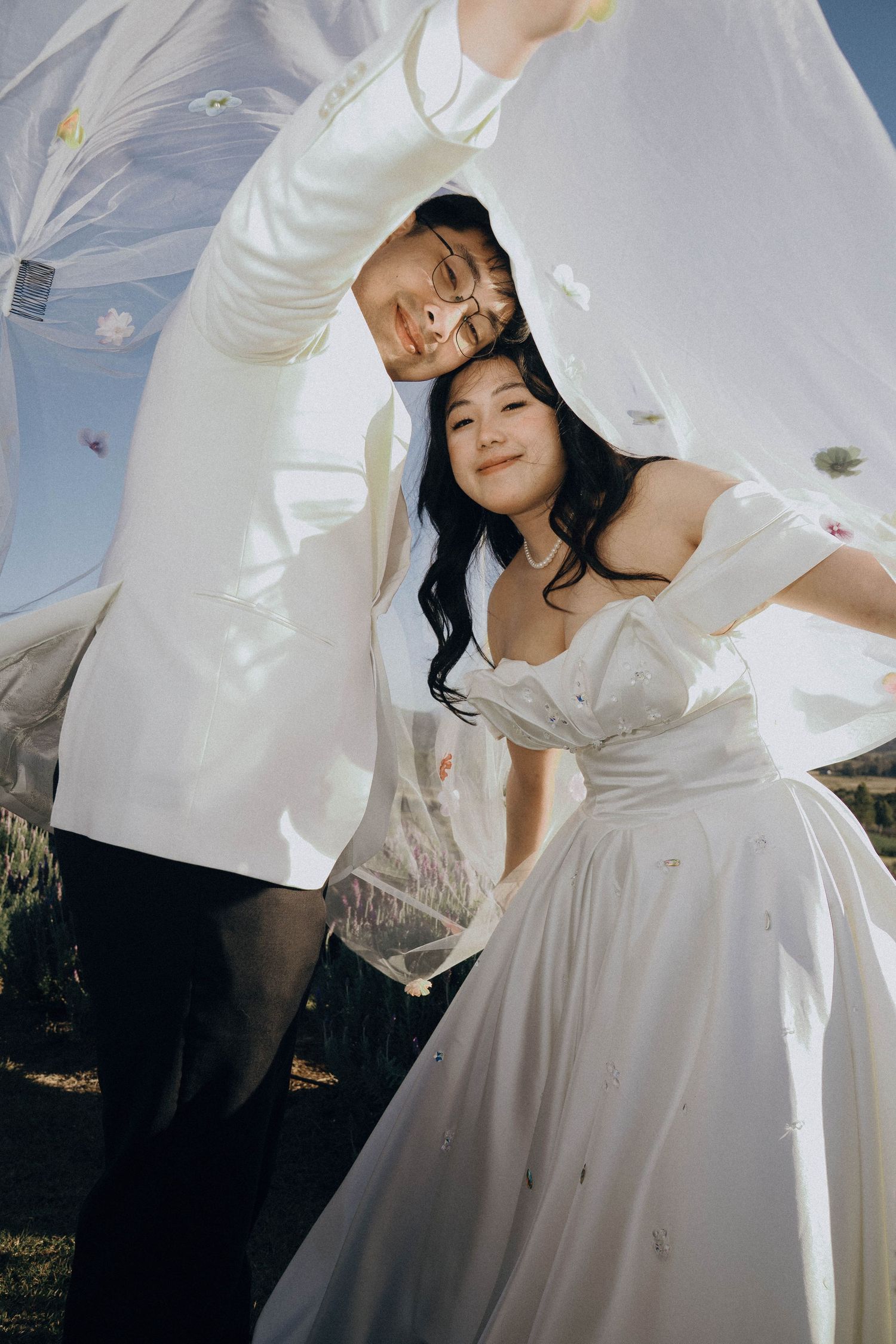 A couple in white wedding attire poses playfully under a veil outdoors during a sunny day.
