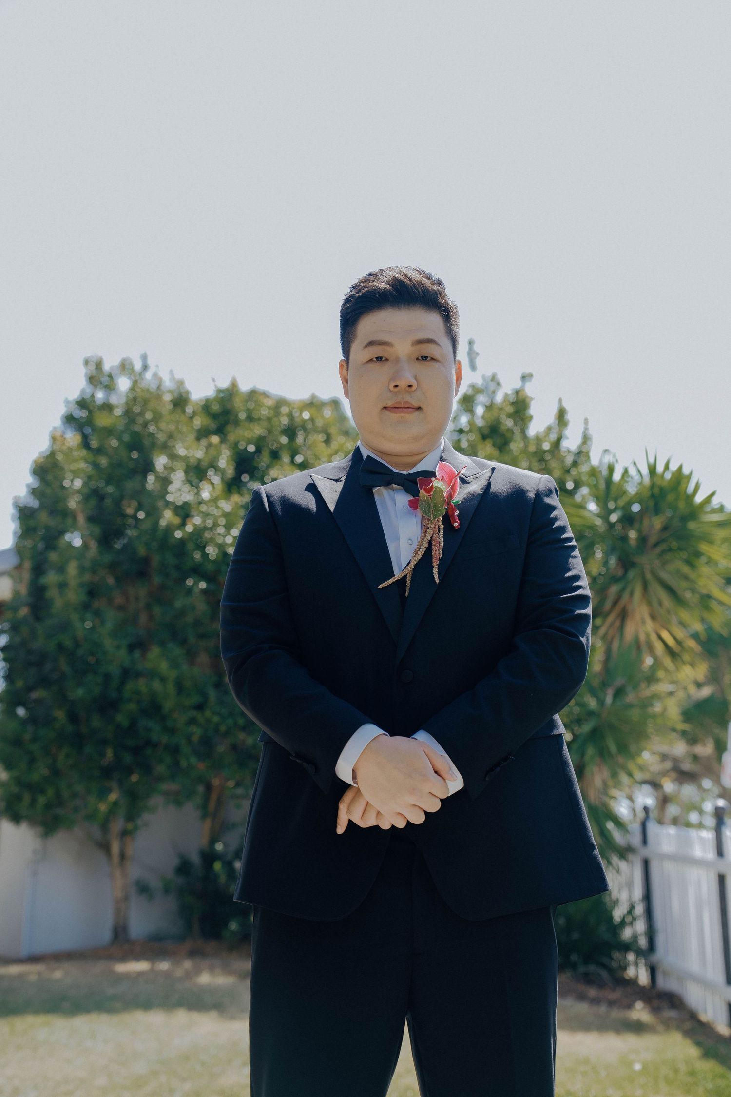 A groom in a classic black tuxedo with a boutonniere stands outdoors on a sunny day.