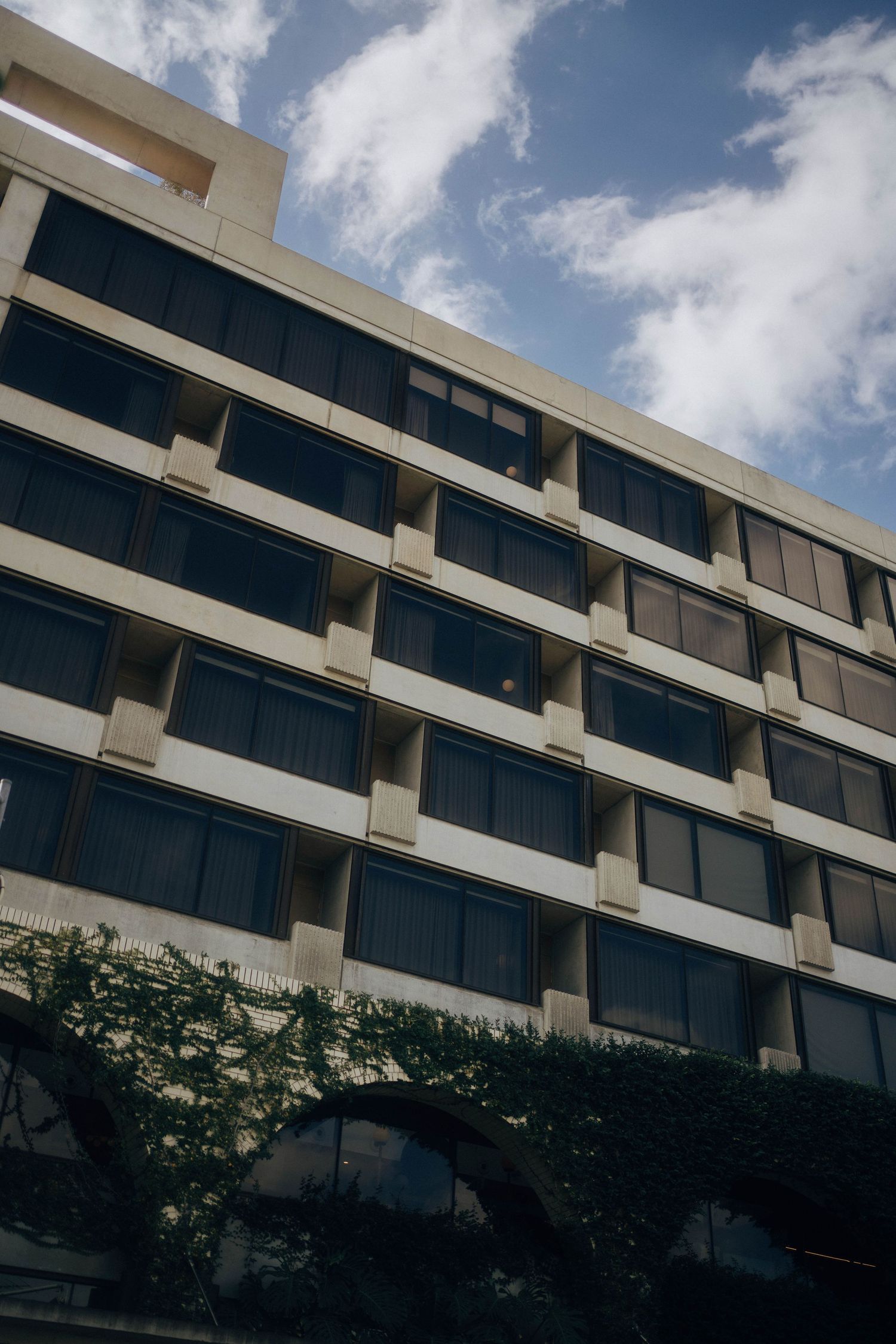 Modern office building with glass windows and concrete facade against blue sky with white clouds and climbing vines below.