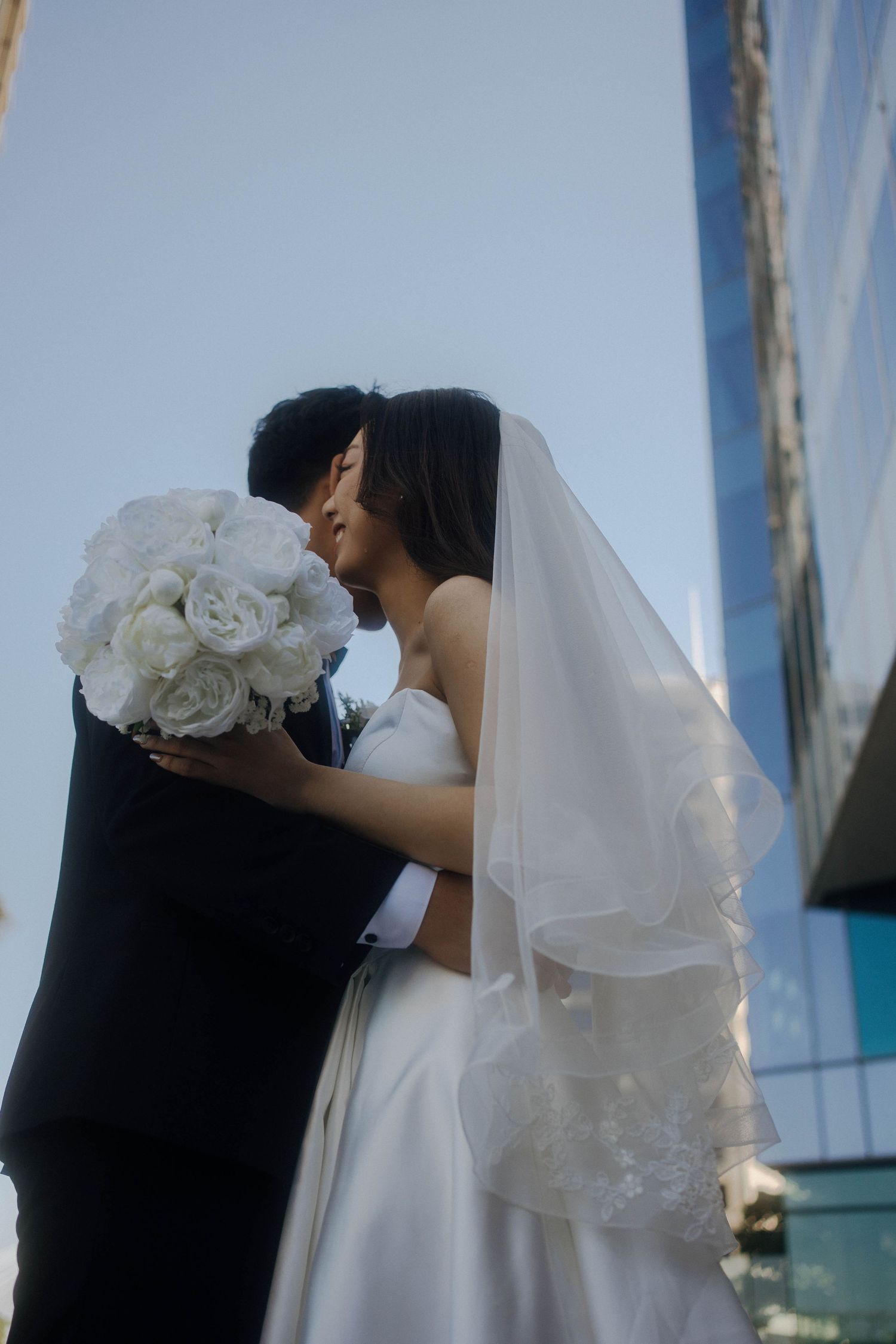 A romantic wedding couple shares a tender kiss against a bright blue sky backdrop.