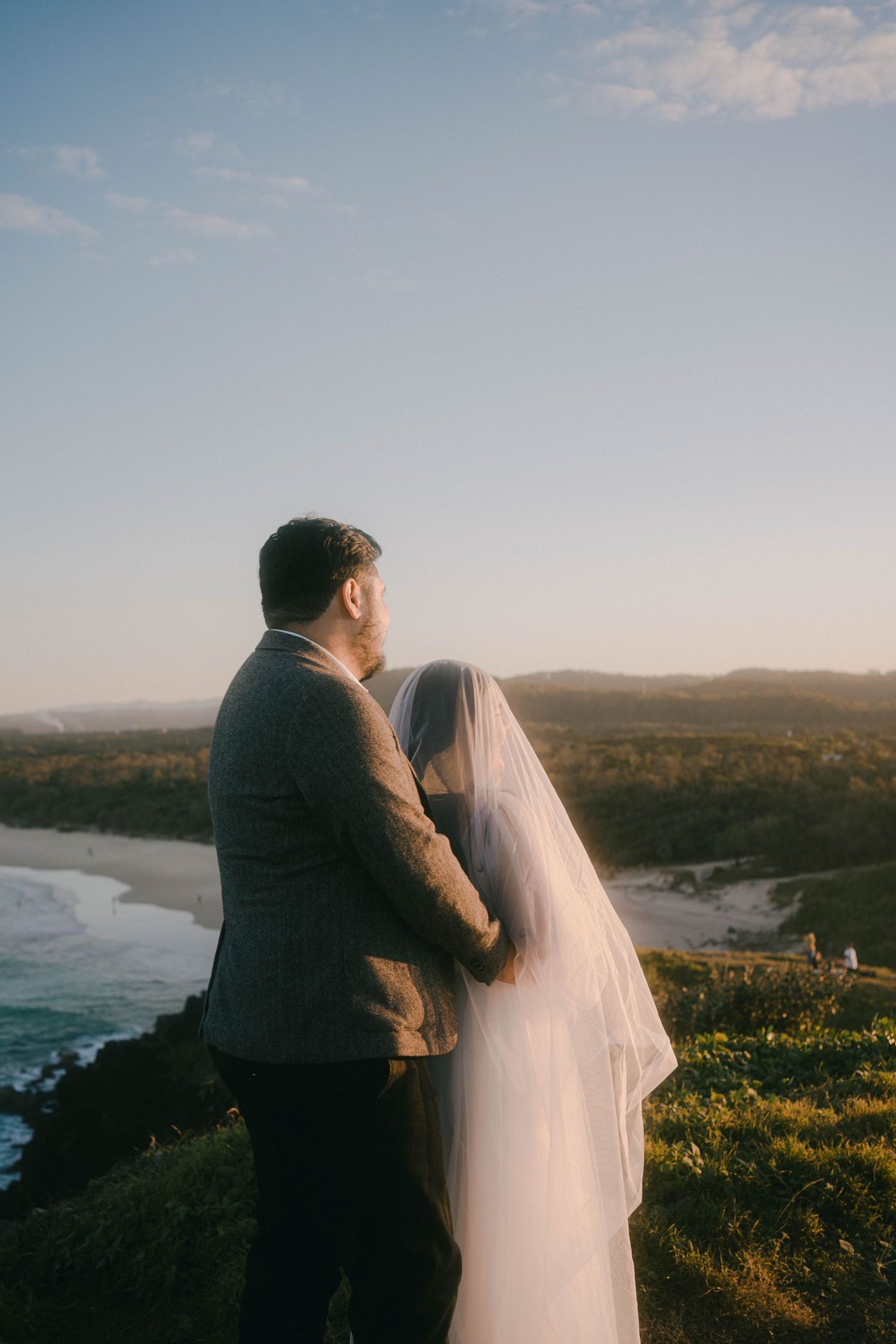 A couple in wedding attire embrace while overlooking a scenic coastal landscape at sunset.