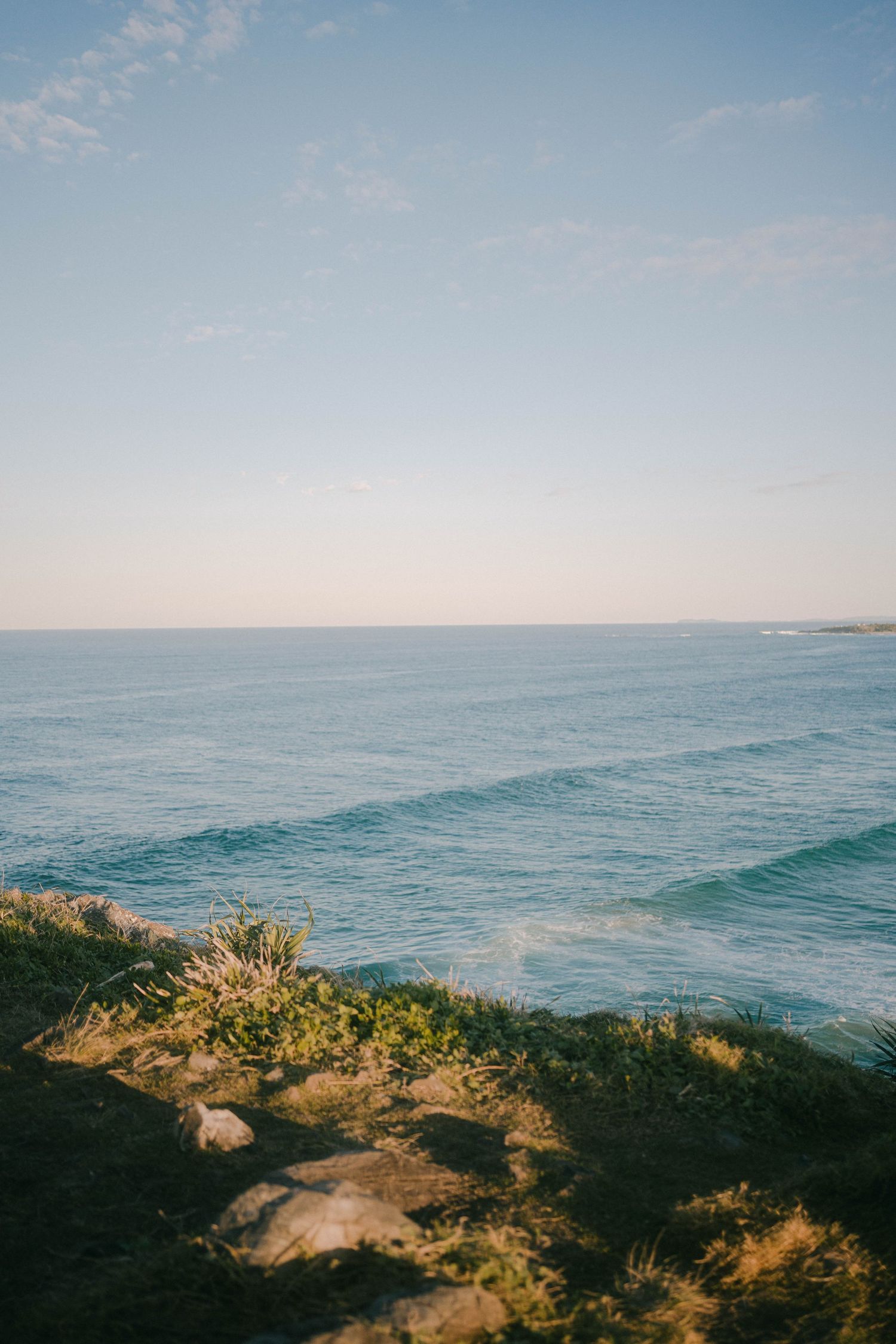 Panoramic coastal landscape showing rocky cliffs with vegetation overlooking the blue ocean horizon at sunset.