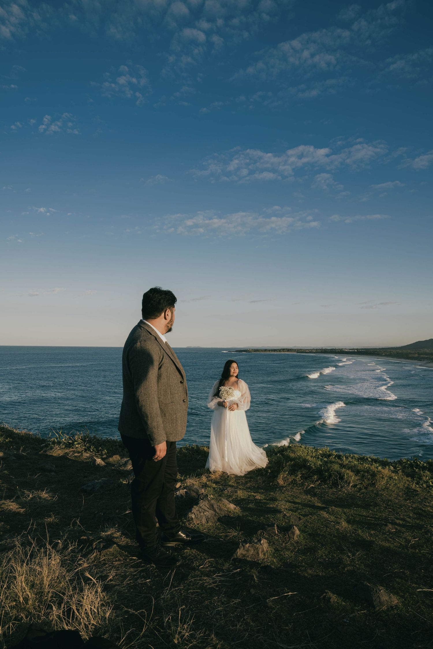 A couple stands on a coastal cliff overlooking the ocean waves during a scenic outdoor wedding photoshoot.