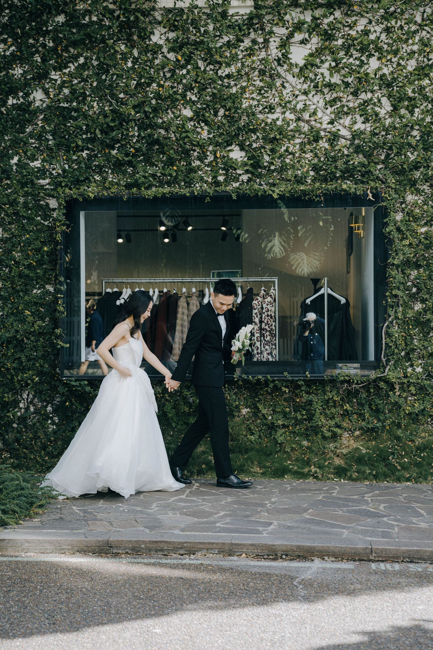 A wedding couple walks together along a stone pathway surrounded by lush greenery and modern architecture.