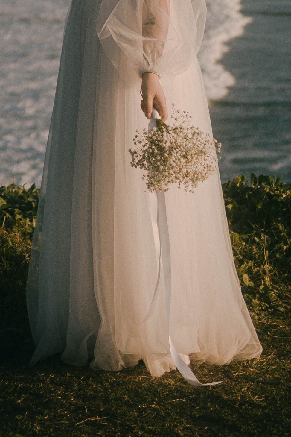 The bottom portion of a flowing white wedding dress with delicate tulle fabric is captured against a natural outdoor setting.