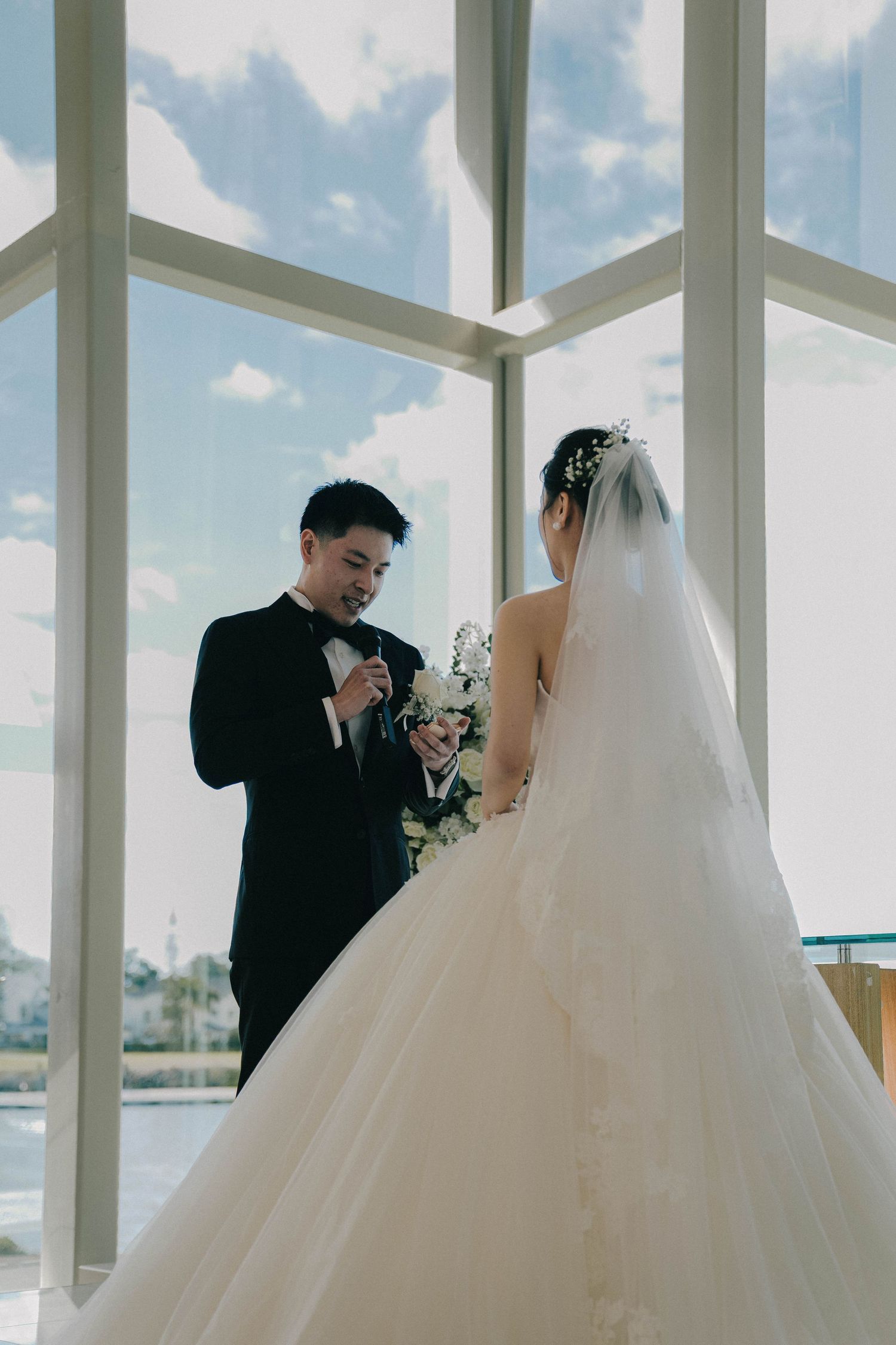 Wedding ceremony in a bright sunlit venue with white decor and large windows overlooking water.