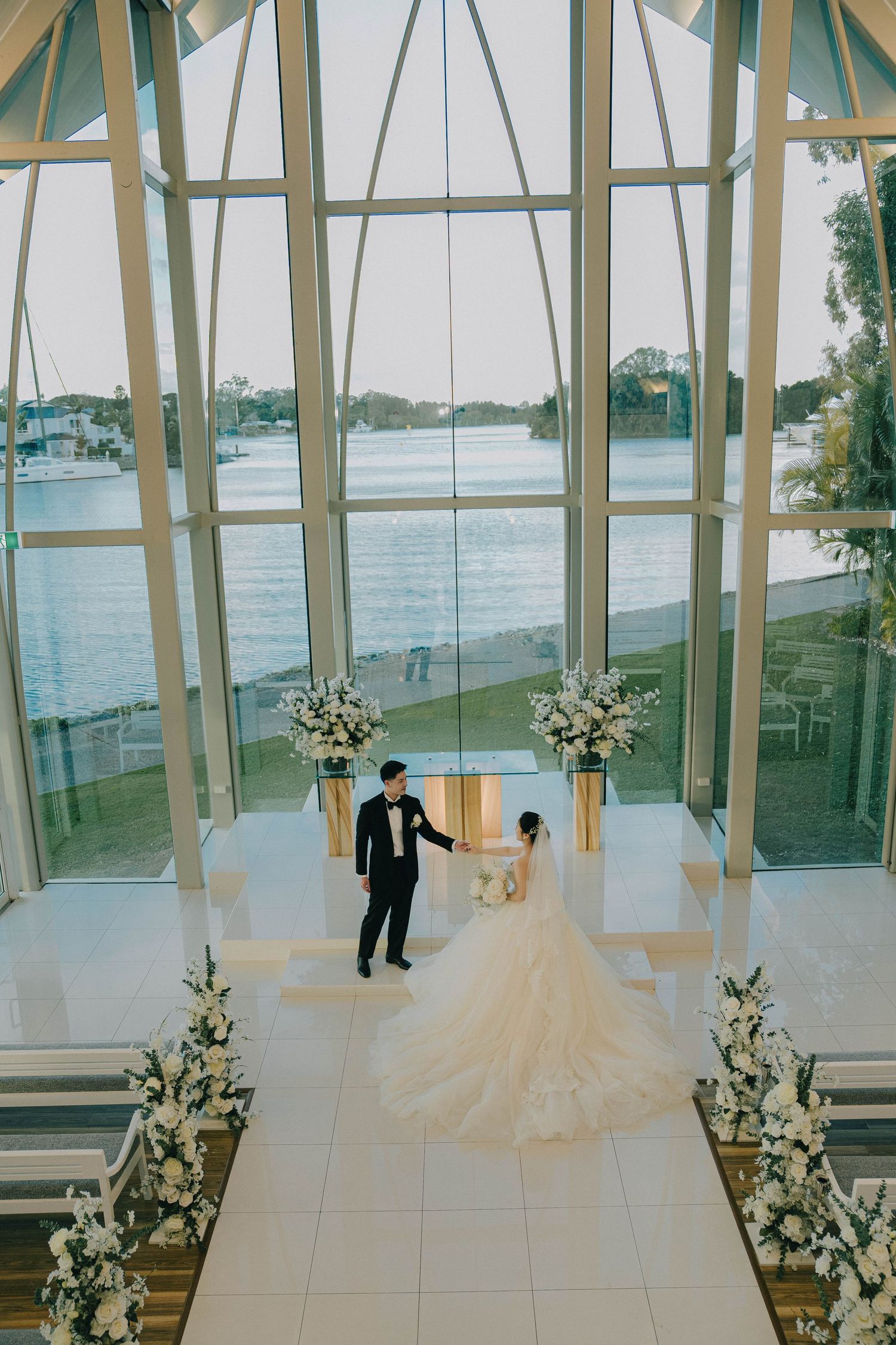 Modern wedding ceremony setup in a bright glass-walled venue with tall windows overlooking a waterfront view.