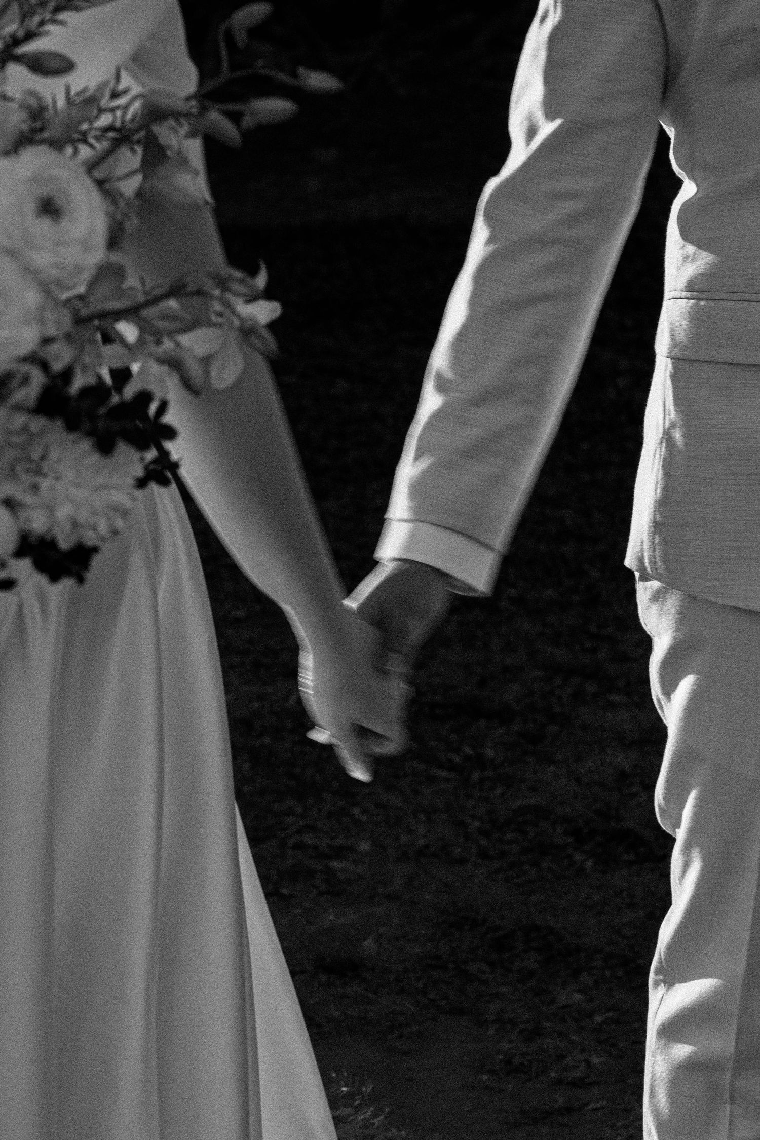 Black and white close-up of newlyweds holding hands, showing white dress and light-colored suit details.
